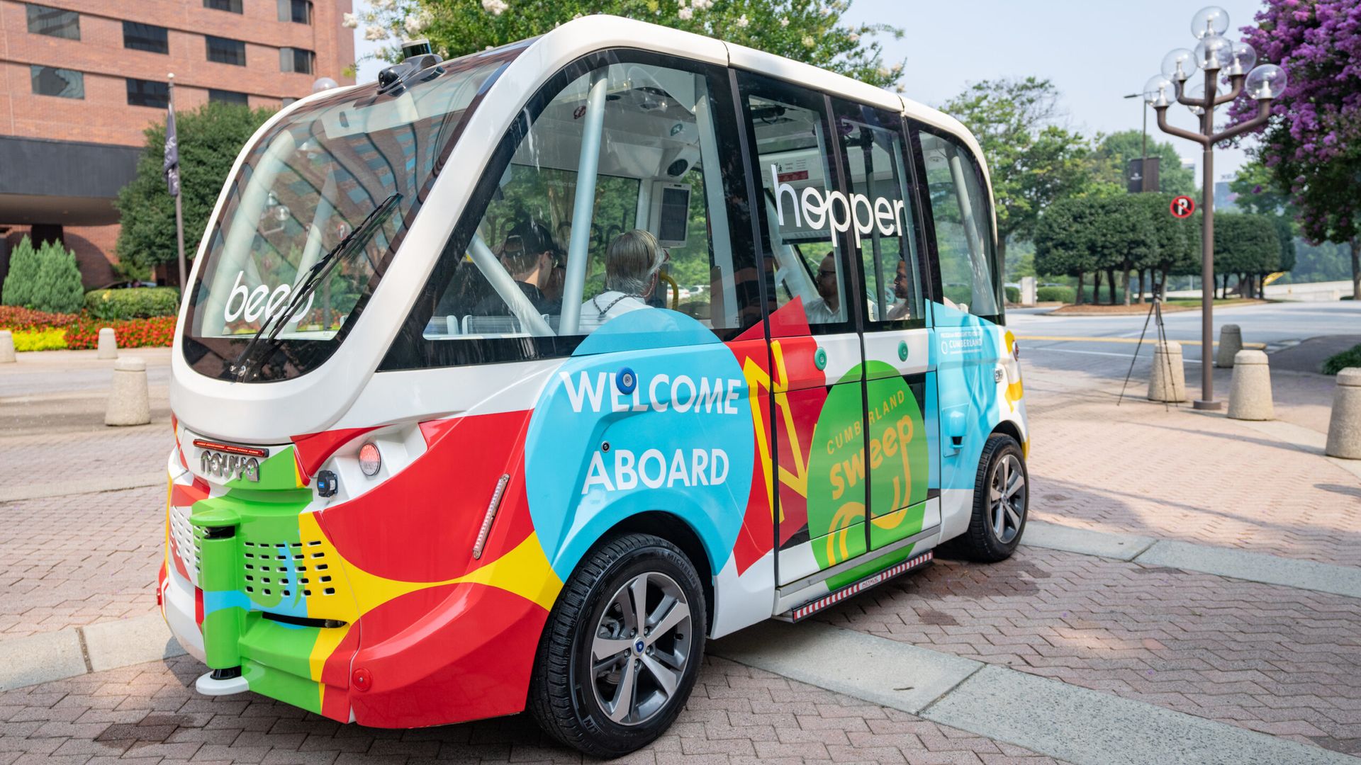 A brightly colored 8-person autonomous shuttle with "Welcome aboard" and "hopper" on the side