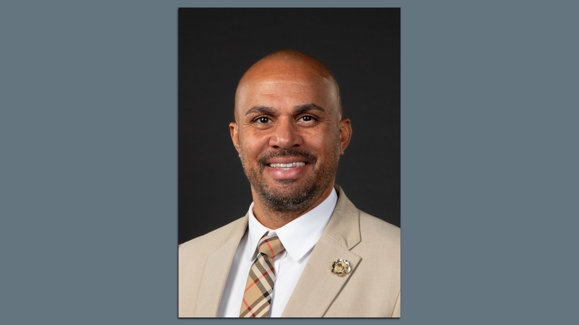 Smiling man with a shaved head in a tan suit and plaid tie, wearing a lapel pin, posed against a dark studio backdrop.