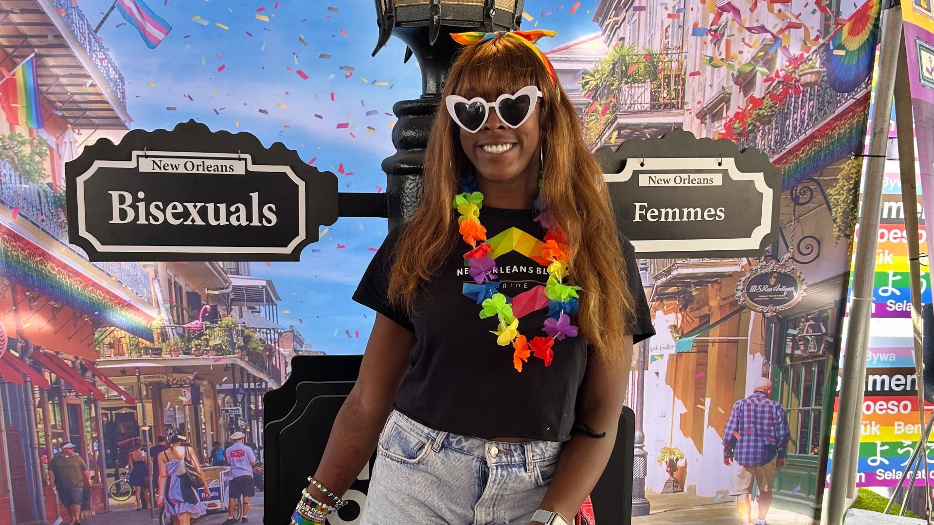 Person with long brown hair wearing heart-shaped white sunglasses, rainbow lei, black New Orleans Pride shirt, and light blue jeans smiles in front of signs reading "New Orleans Bisexuals" and "New Orleans Femmes".