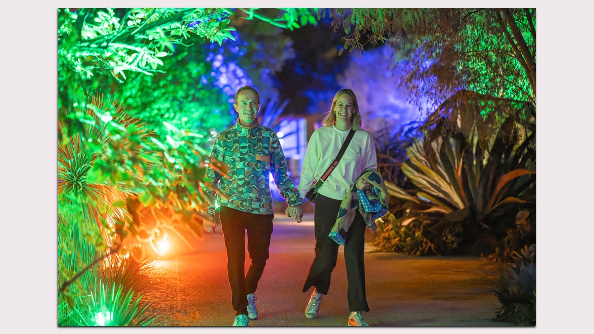 A couple holding hands and smiling while walking on a pathway surrounded by green, orange, and blue illuminated plants at night.