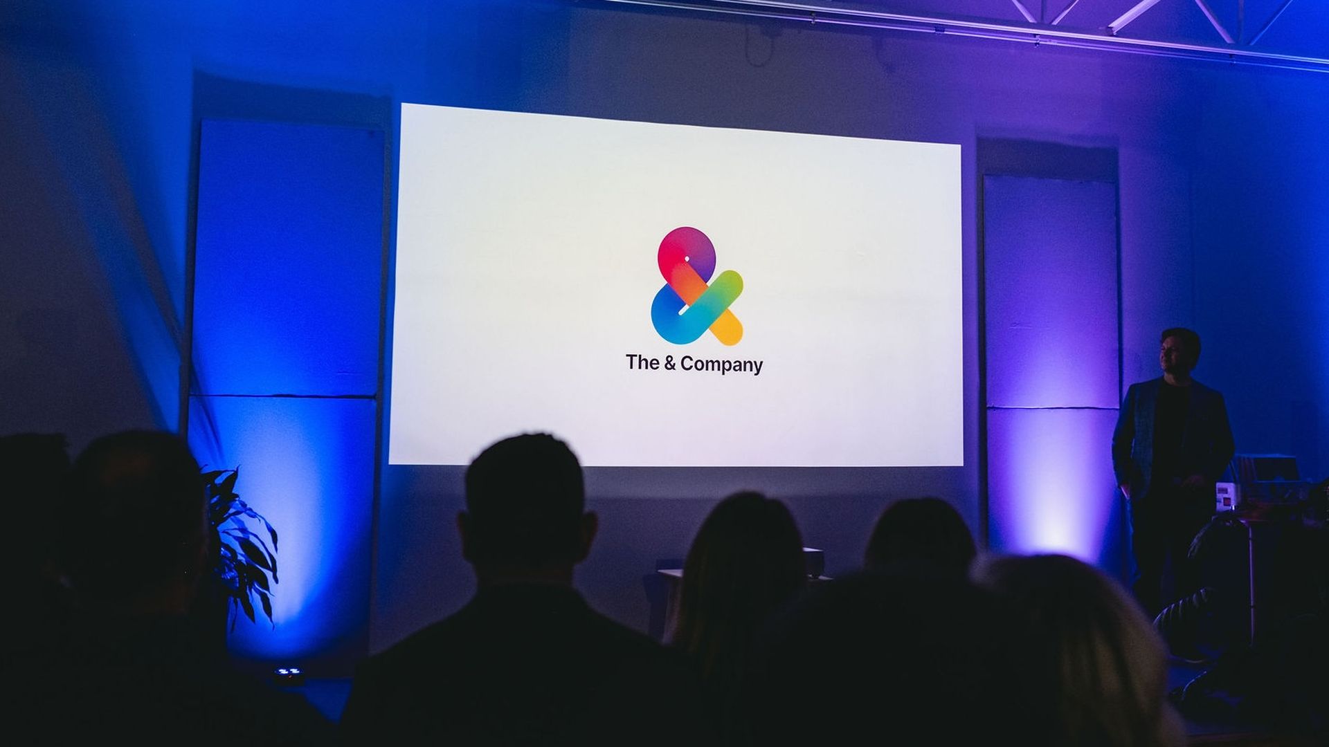 A dark room with blue and purple lighting shows a large white screen displaying a colorful ampersand logo above the text "The & Company" as an audience watches a presenter.