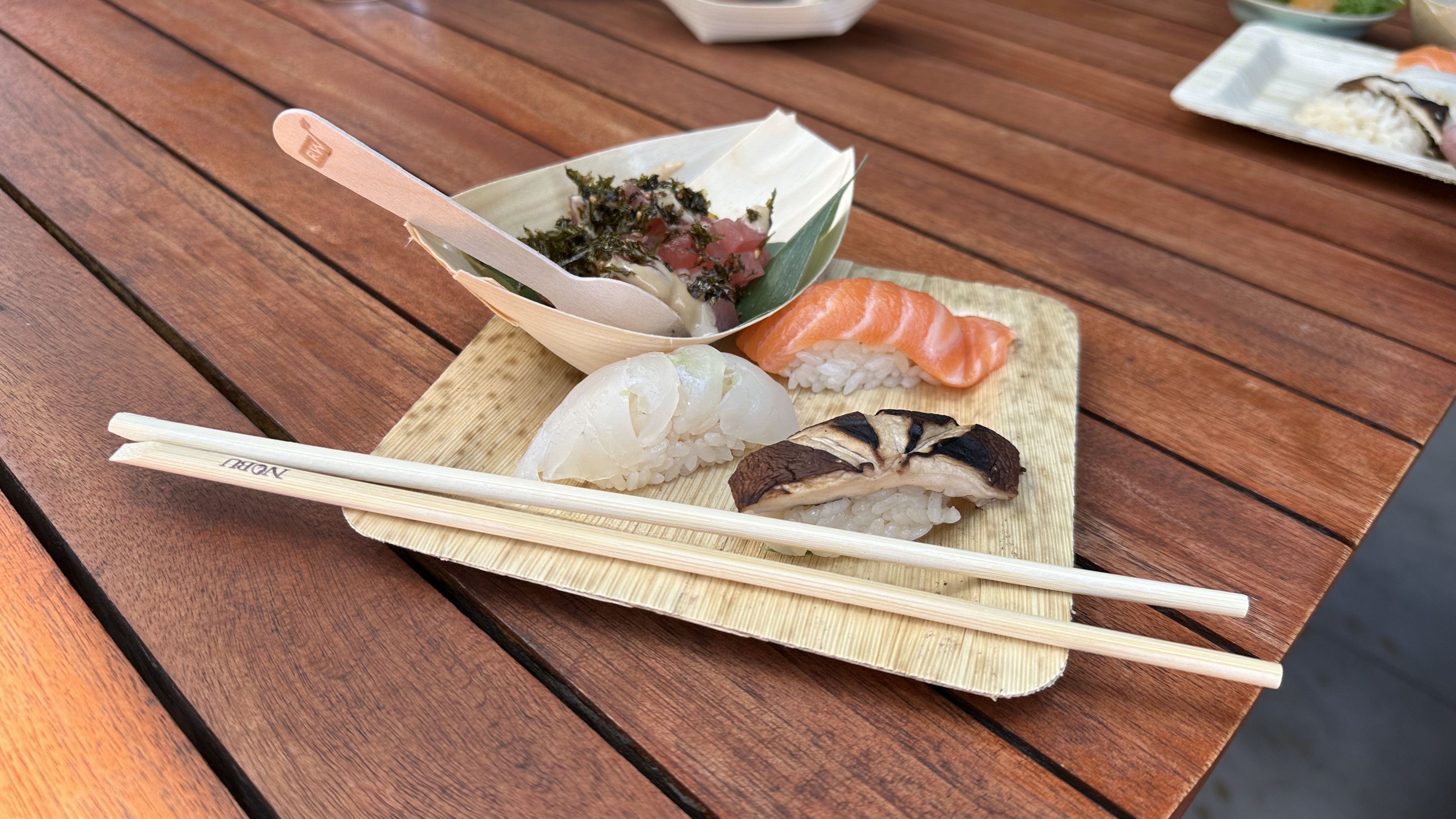 A small bamboo plate has three pieces of sushi and a poke bowl sitting on it on a wooden tabletop with a pair of Nobu-branded chopsticks.