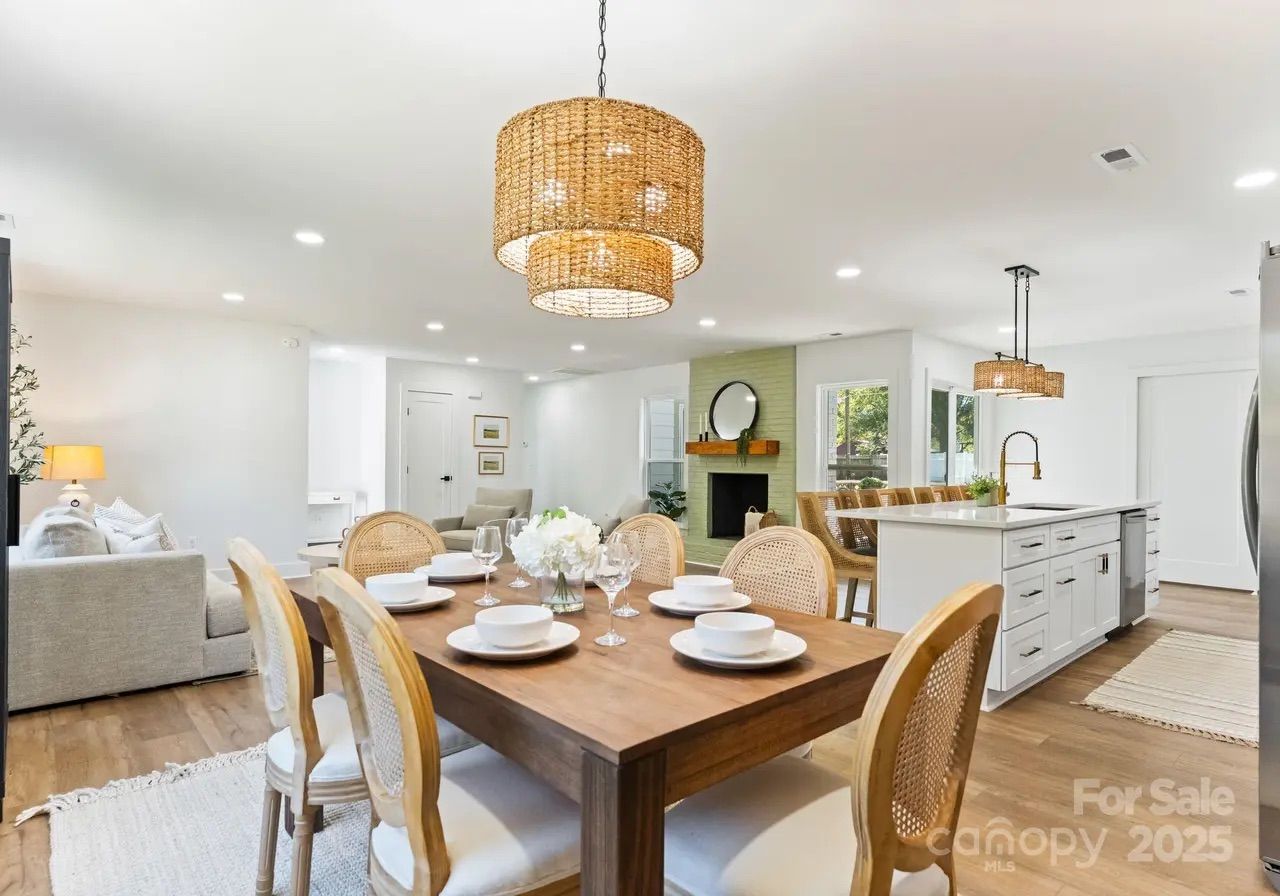 Bright open dining and kitchen area with wooden table set with white dishes, woven chairs, large wicker pendant lights, light walls, and hardwood floors.