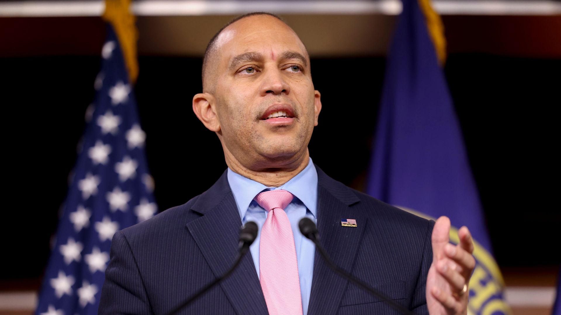 House Minority Leader Hakeem Jeffries, wearing a blue pinstripe suit and speaking into microphones in front of a pair of flags.