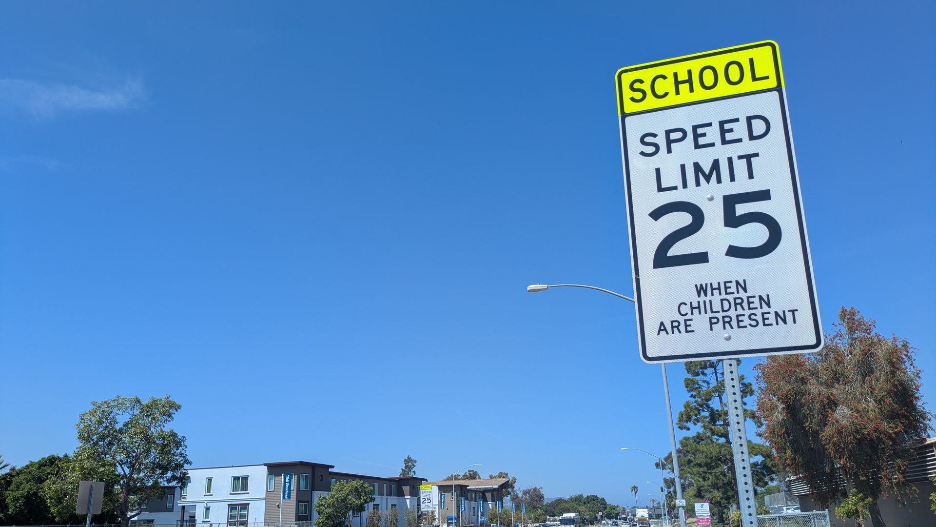 Bright blue sky over a suburban street; a tall, angled school sign displays speed limit 25 when children are present; cars line the road and a sidewalk runs along the right.