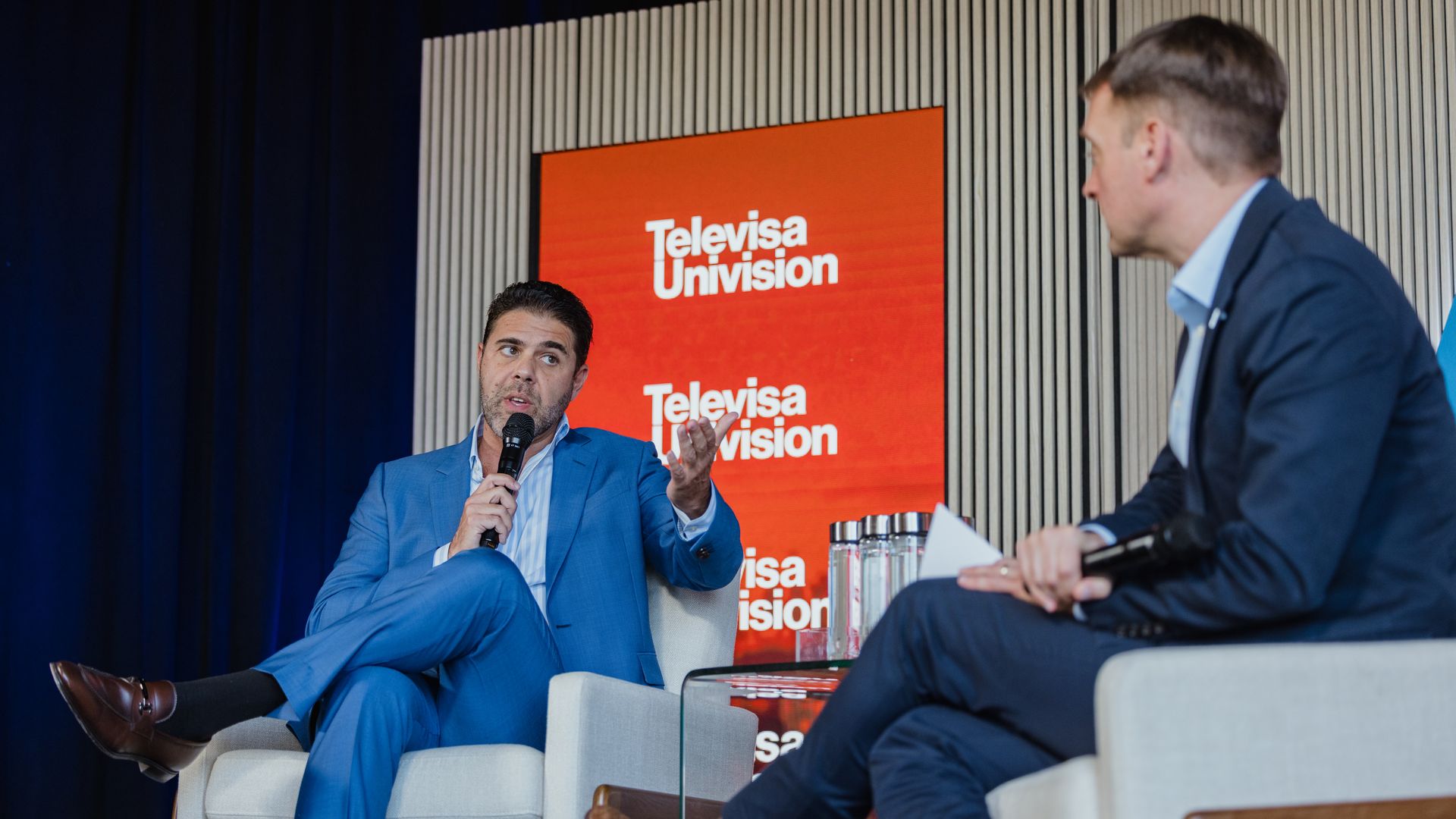 Two men in suits on stage; the left speaks into a microphone and gestures. A red Televisa Univision display backs them, with water bottles on a glass table between white chairs.