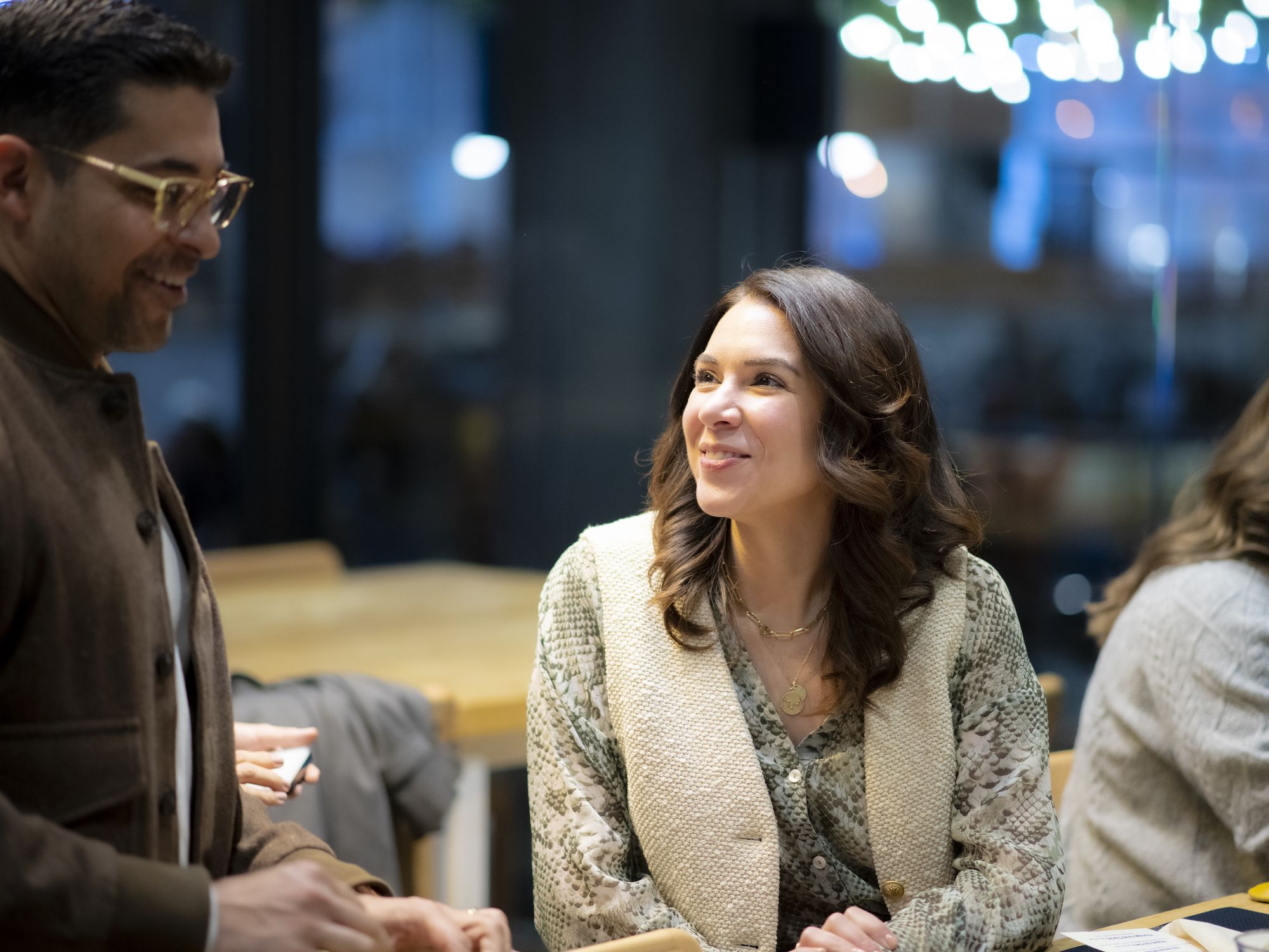 Smiling woman with wavy brown hair wearing a patterned shirt and cream vest, sitting at a table in a warmly lit modern indoor space, conversing with a man in glasses and brown jacket.