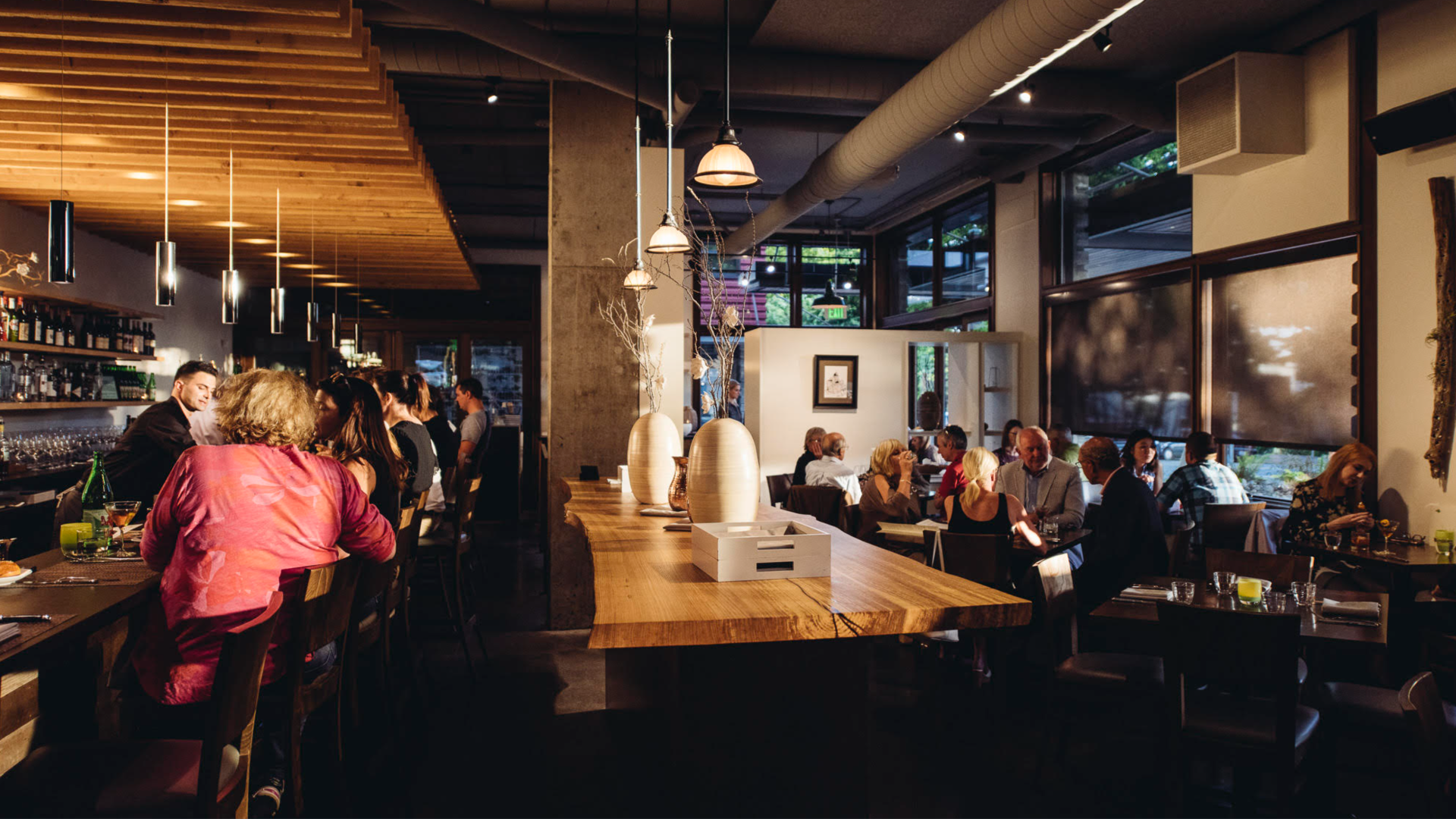 People sit at a bar and at tables Copine. The dimly lit restaurant space has pendant lighting and wooden accents.