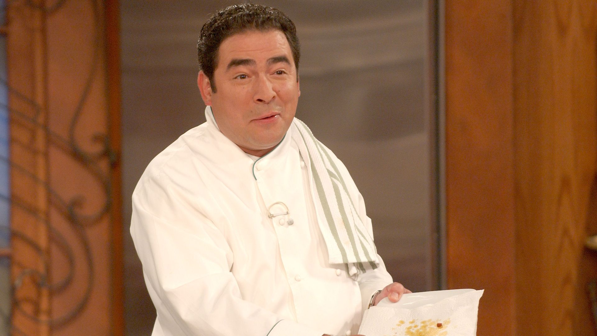 Emeril Lagasse, wearing a white chef's coat, tilts an oily plate toward the camera.