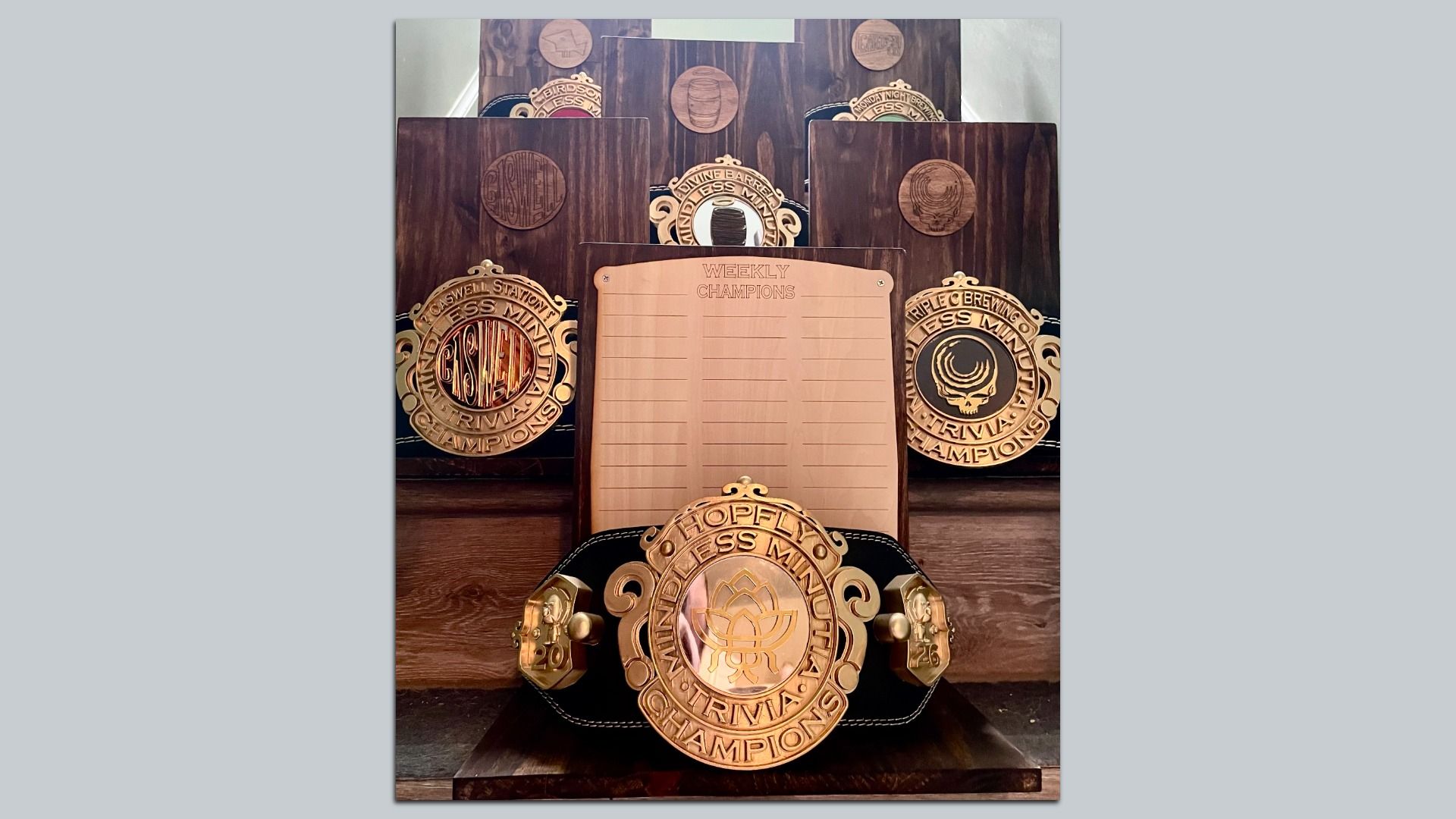 Display of dark wooden plaques with circular medallions surrounding a central copper board labeled "WEEKLY CHAMPIONS." A large gold championship belt sits at the front with bronze accents and decorative trim.