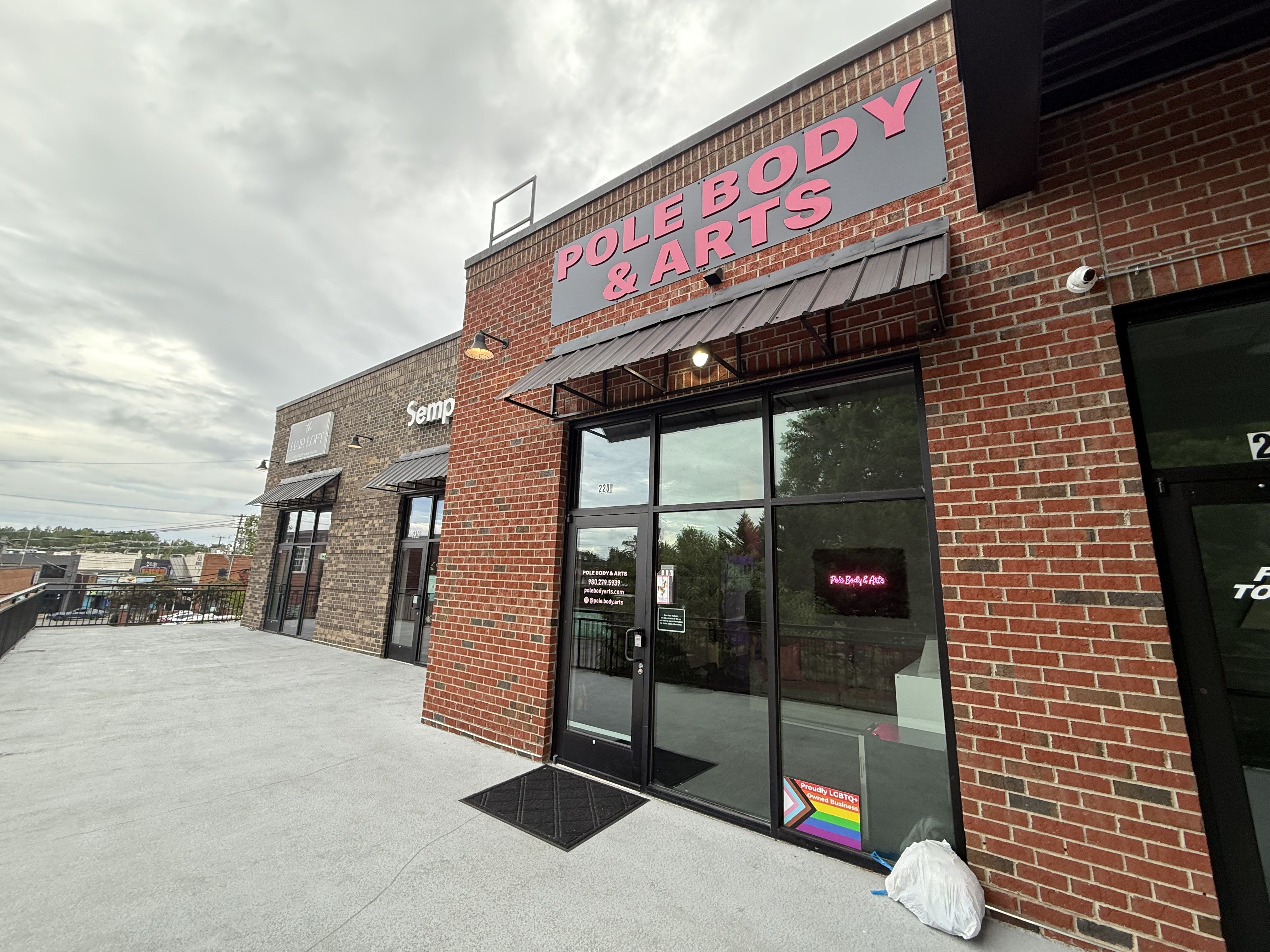 Exterior view of a brick building with a sign in bold pink letters reading "POLE BODY & ARTS" above a glass door and window, cloudy sky overhead and a white bag on the ground.
