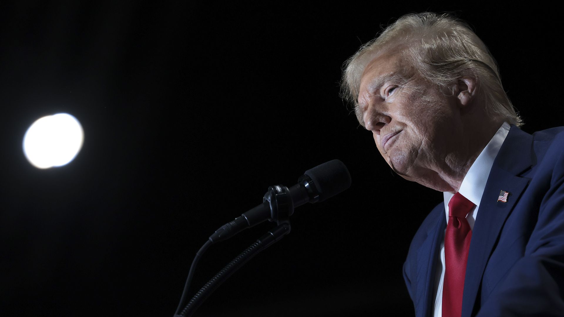 Trump in a blue suit and red tie looking off to the left in front of a black background.