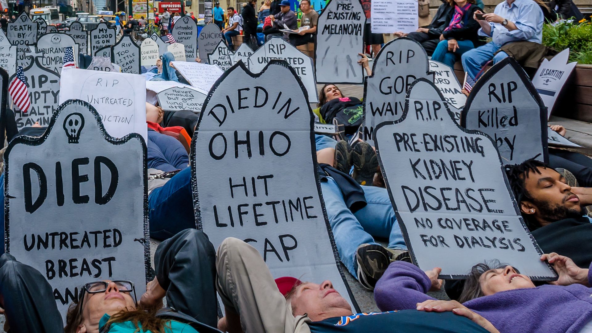Protesters lie in front of gravestones saying "Died of untreated breast cancer" and other slogans