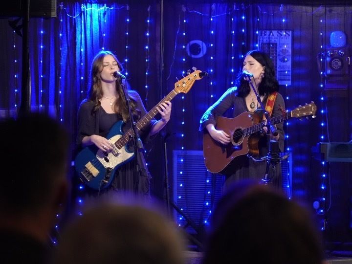 Two women holding guitars sing into microphones on a small stage with blue string lights hanging vertically in the background. 