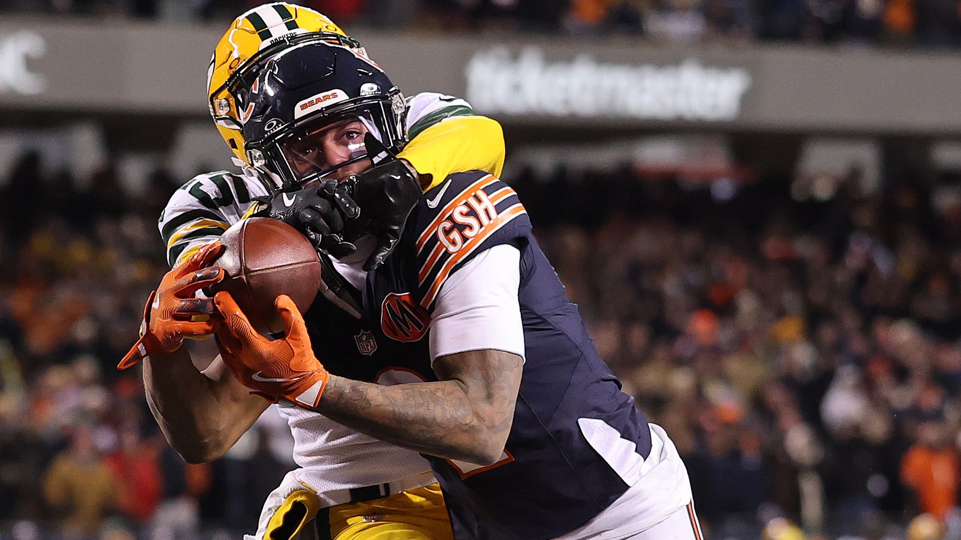 NFL player in a navy Bears jersey with orange gloves catching a football while a defender in a green and yellow Packers uniform tackles him from behind during a game.