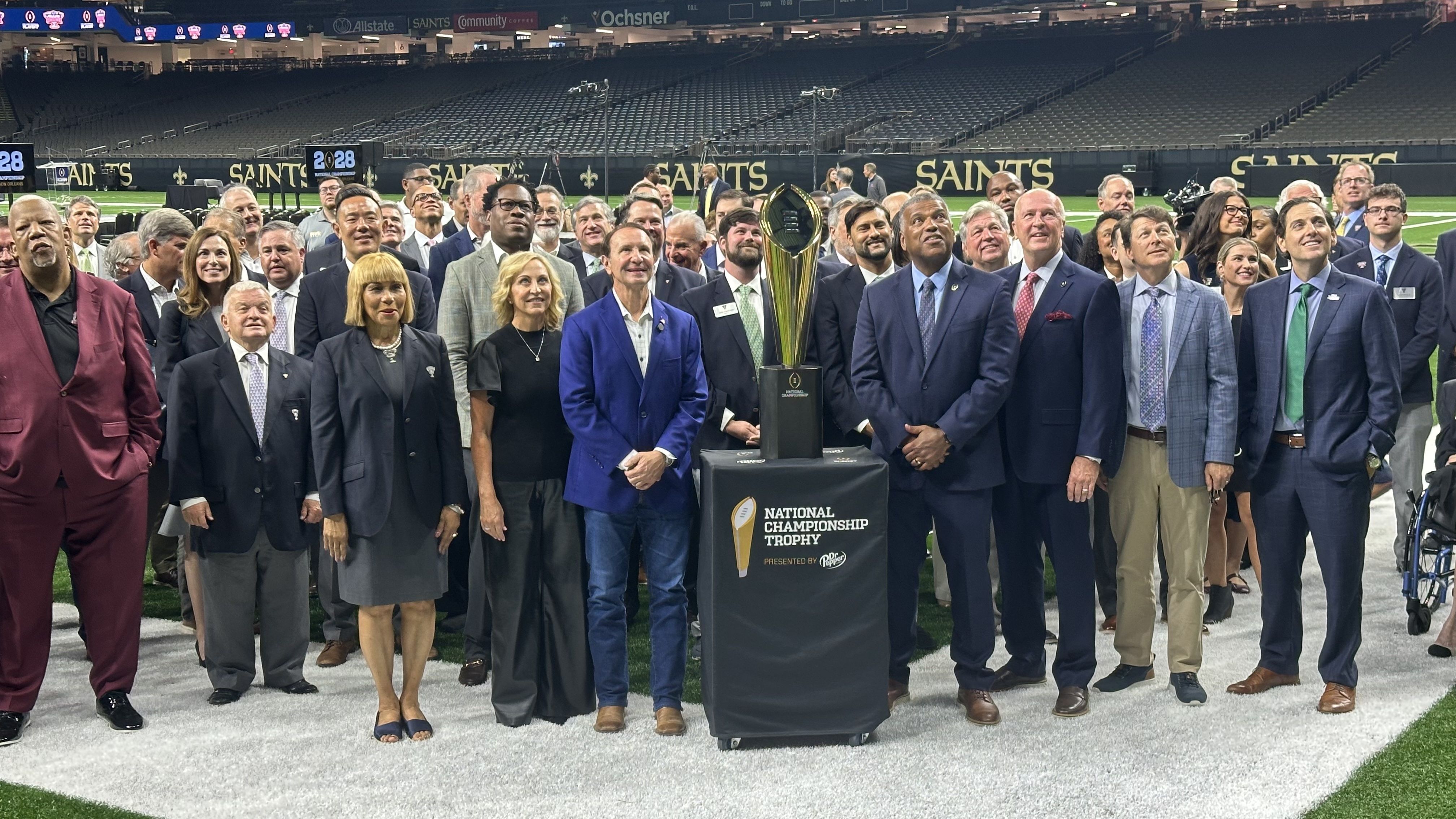 A large group of people, mostly in formal attire, stand on a football field behind the National Championship Trophy on a pedestal with Saints stadium seats and signage in the background.