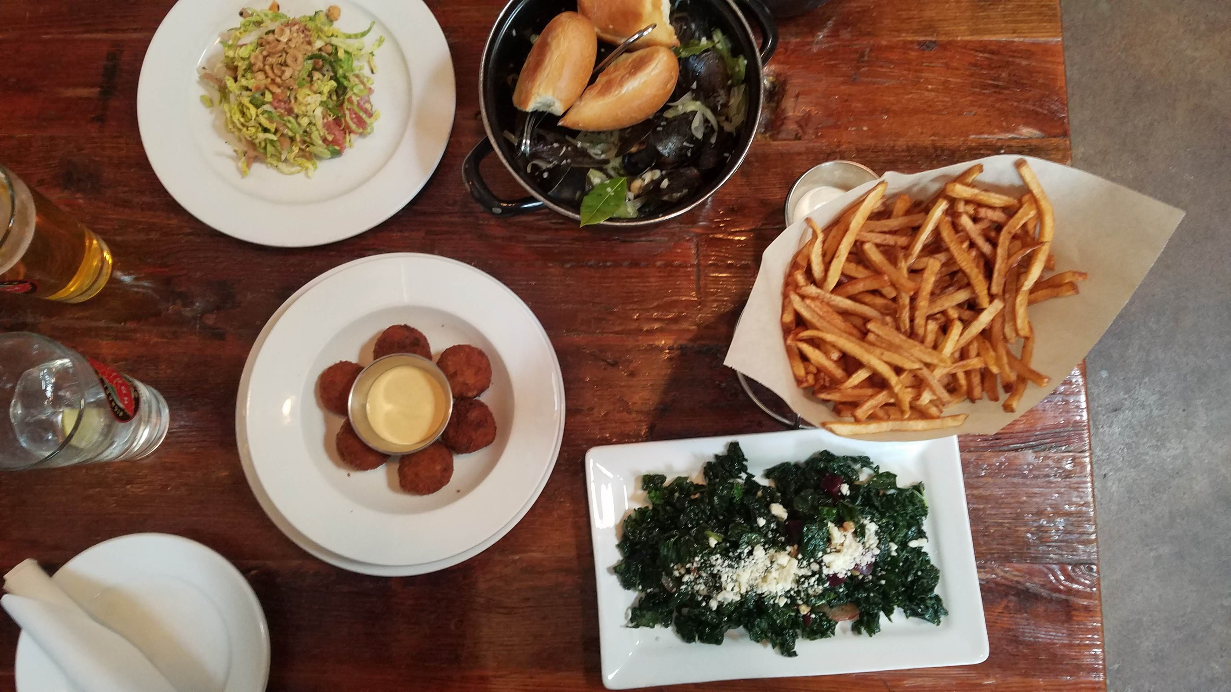 Overhead view of a wooden table with plates of fried croquettes and sauce, a leafy green salad with feta, French fries in a basket, a pot with bread rolls and mussels, and a salad with shredded greens and nuts.