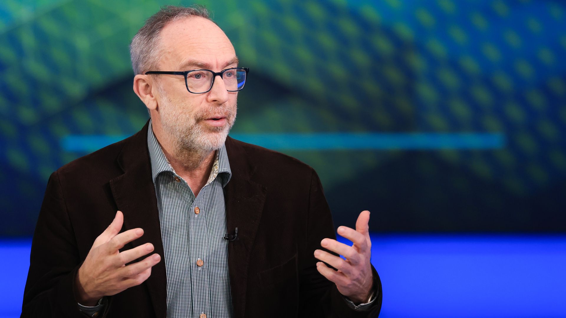 Wikipedia founder Jimmy Wales speaks during a television interview, wearing a dark suit jacket in front of a tv set backdrop.