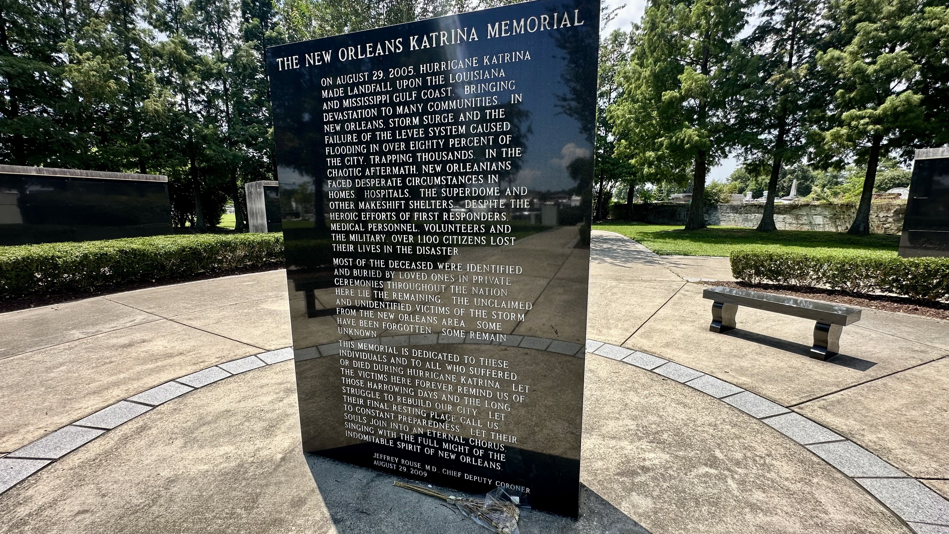 Black polished stone memorial in a park with engraved text about Hurricane Katrina's impact in New Orleans. A bench and trees are visible nearby under a sunny sky.