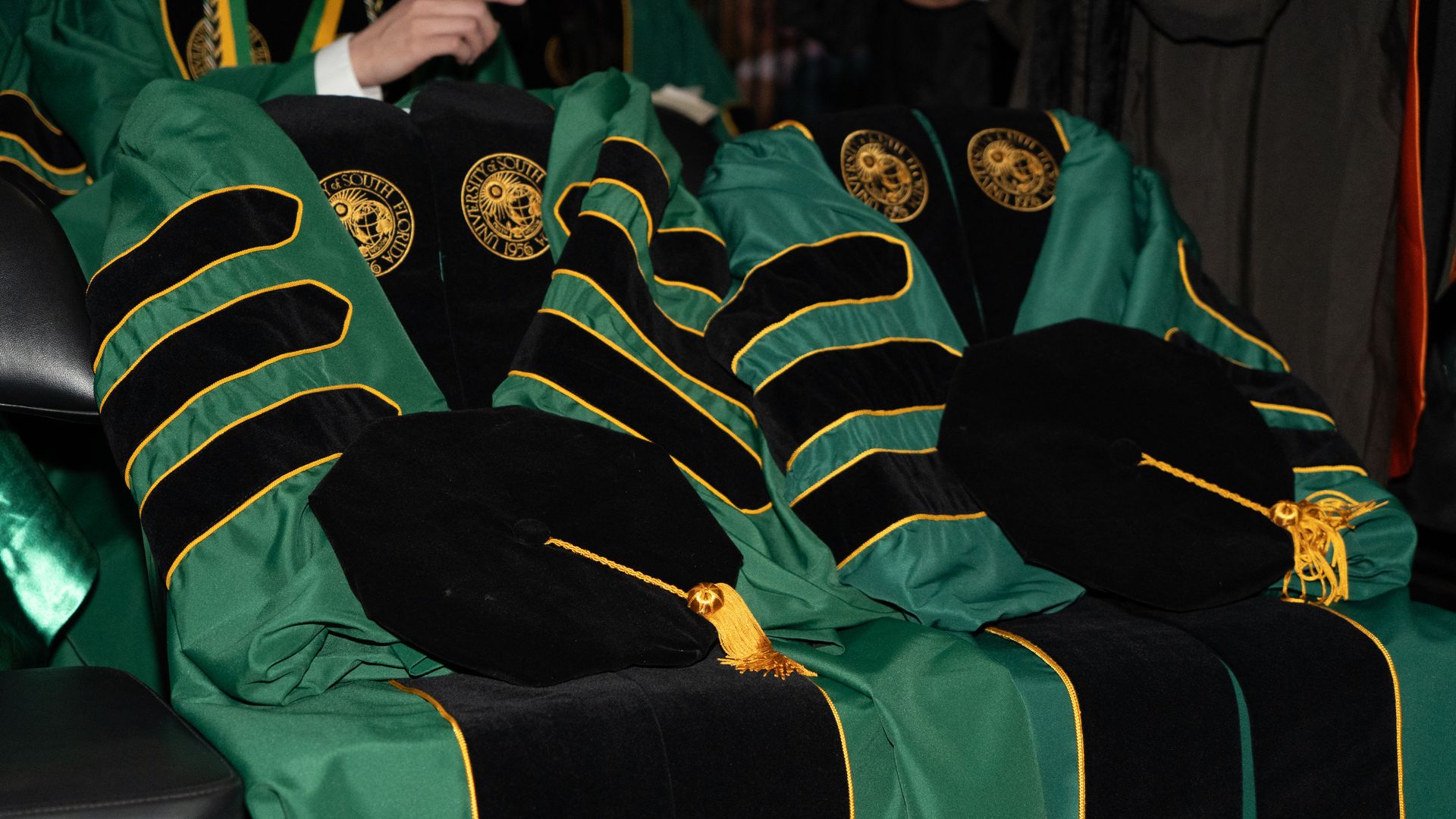 Two green and black graduation gowns with gold trim and tassels, featuring circular gold seals on the fabric. The gowns are empty; no one is wearing them.