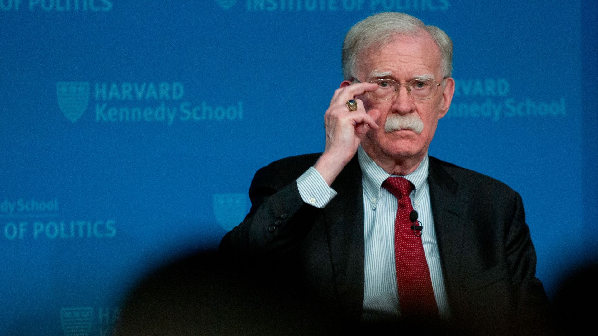 Former US national security adviser,John Bolton, with gray hair and a moustache, wearing a black jacket, blue pin strip suit and red tie, touches the slim, silver frames of his glasses in front of a blue background.
