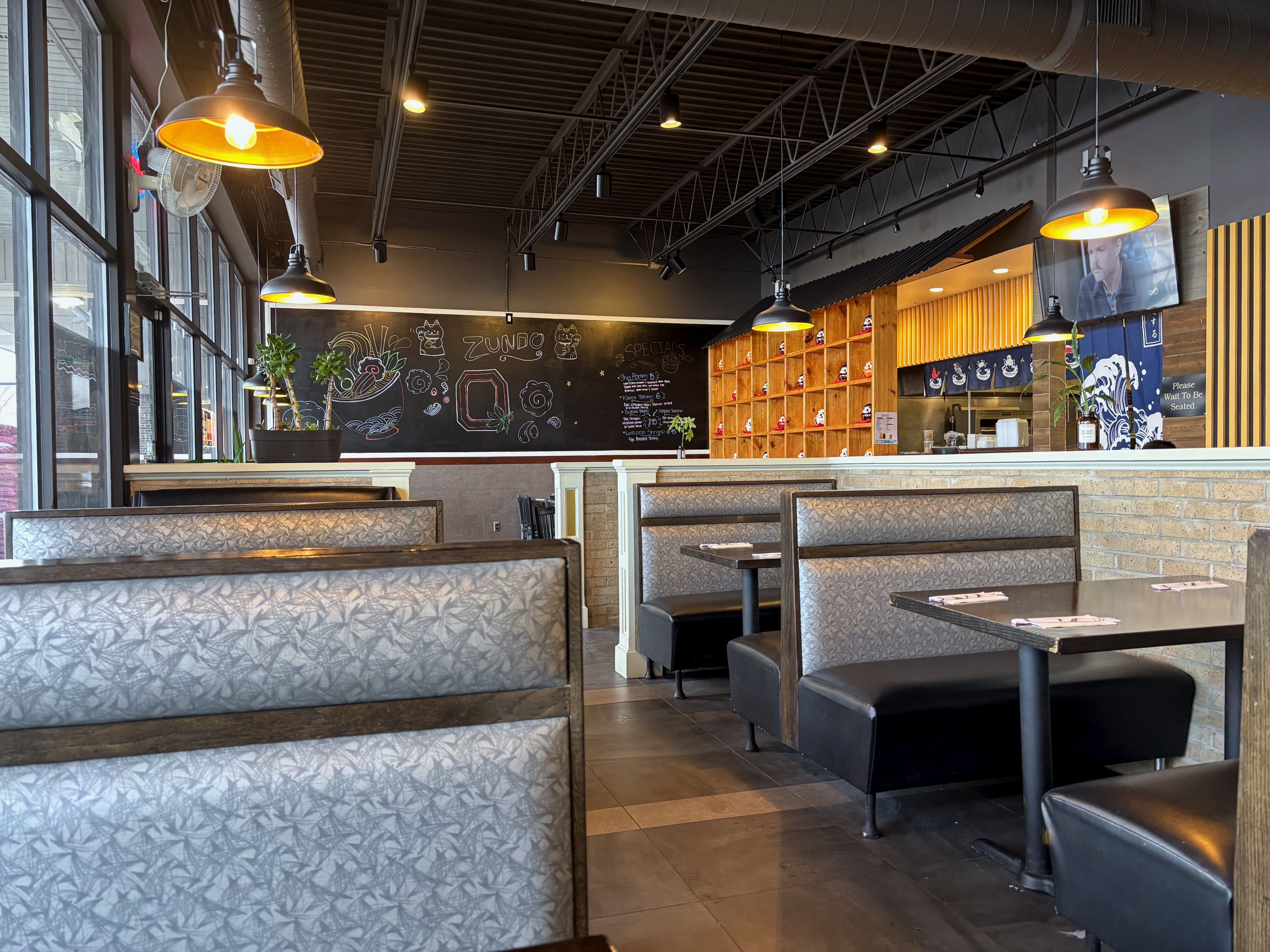 Interior of a cozy restaurant with gray booths, black tables, hanging pendant lights, a chalkboard with colorful drawings and specials, wooden shelves with decor, and large windows.