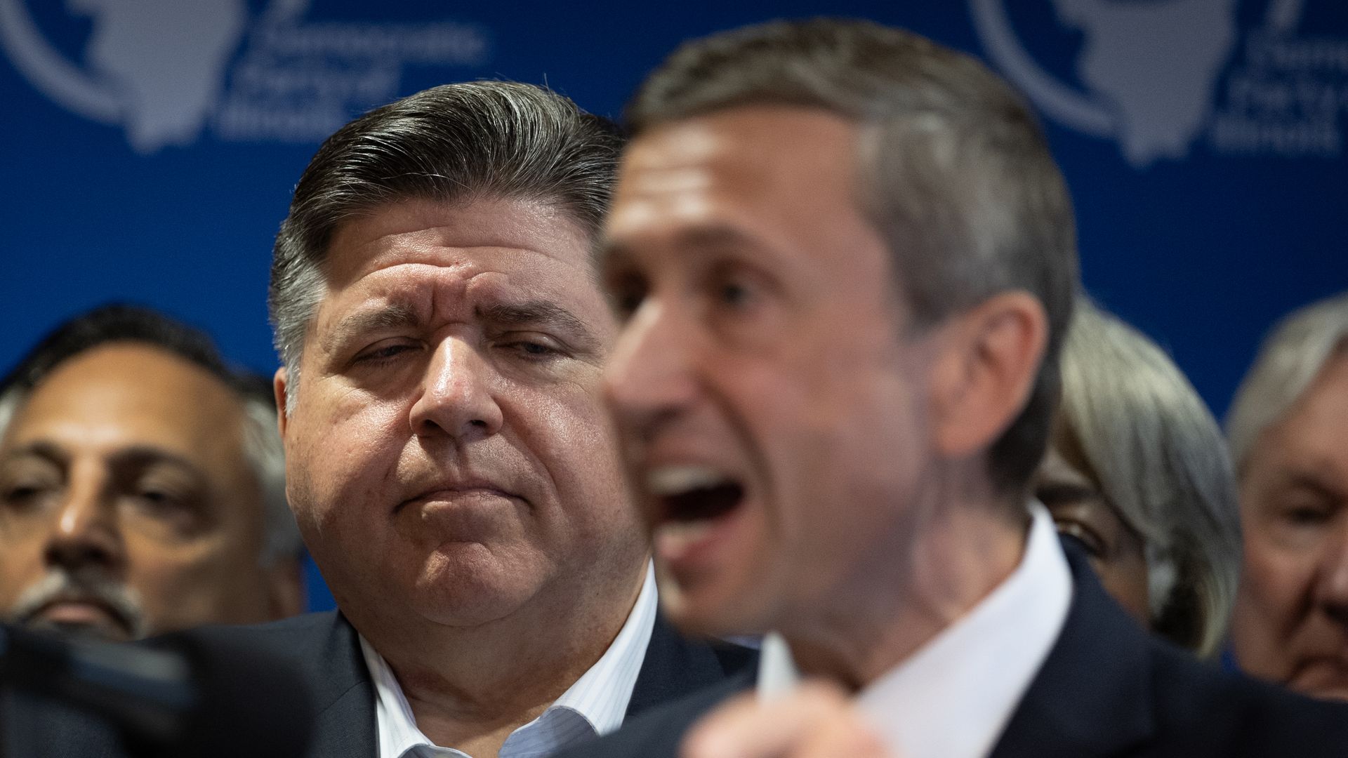 Man in suit with gray-streaked hair frowning and looking down, standing among others at a press event with blue background and world map logos.
