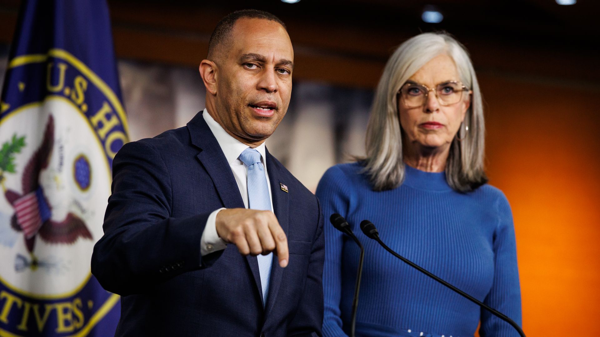 Two political figures at a podium: a man in a navy suit and light blue tie speaks, while a woman in a blue dress stands beside him; a flag and official seal are visible in the background.