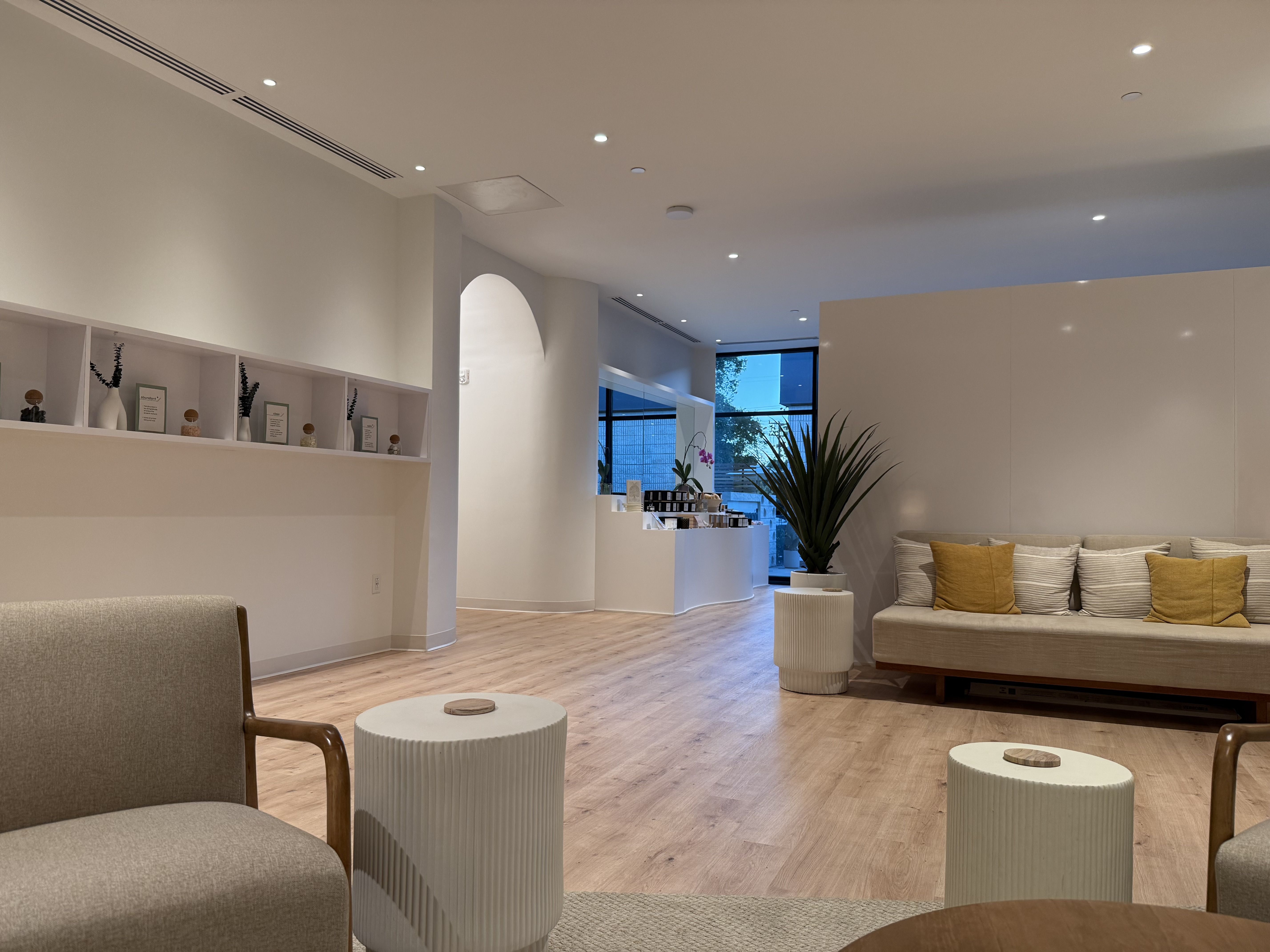 Modern, airy lobby with pale walls, wooden floor, and a large window. Left shelves hold small decor; a curved arch leads to seating with beige cushions and mustard-yellow pillows.