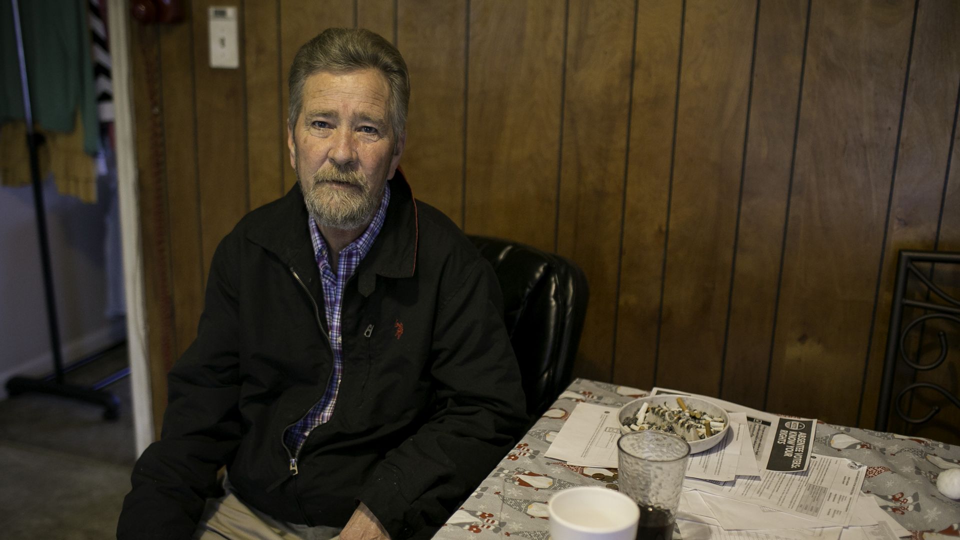 photo of a man named McCrae Dowless sitting at a kitchen table with wood paneling behind him and cigarettes in an ashtray