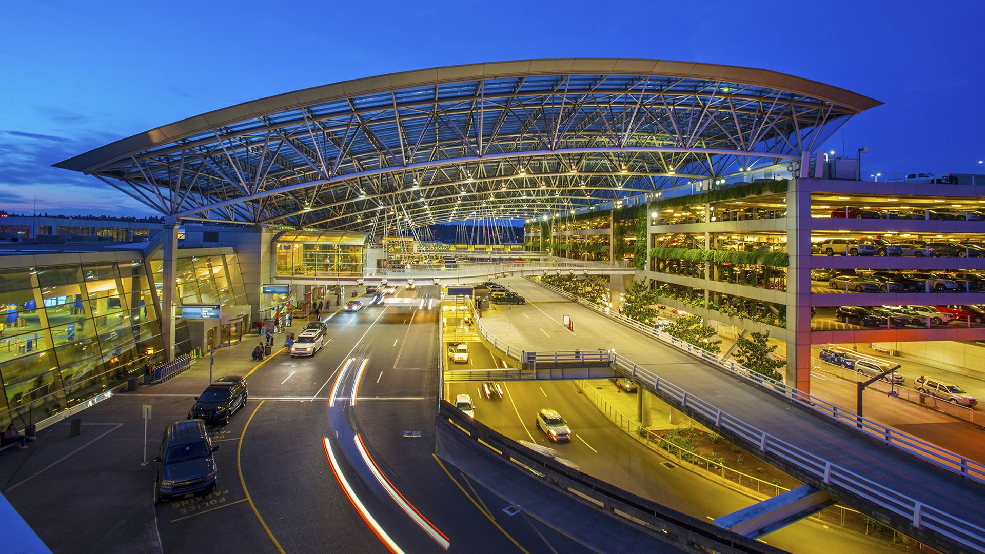 Twilight view of a modern airport terminal under a deep blue sky, featuring a large curved glass and steel roof, lit concourse, and a multi-level parking garage to the right; light trails from moving cars on the road.