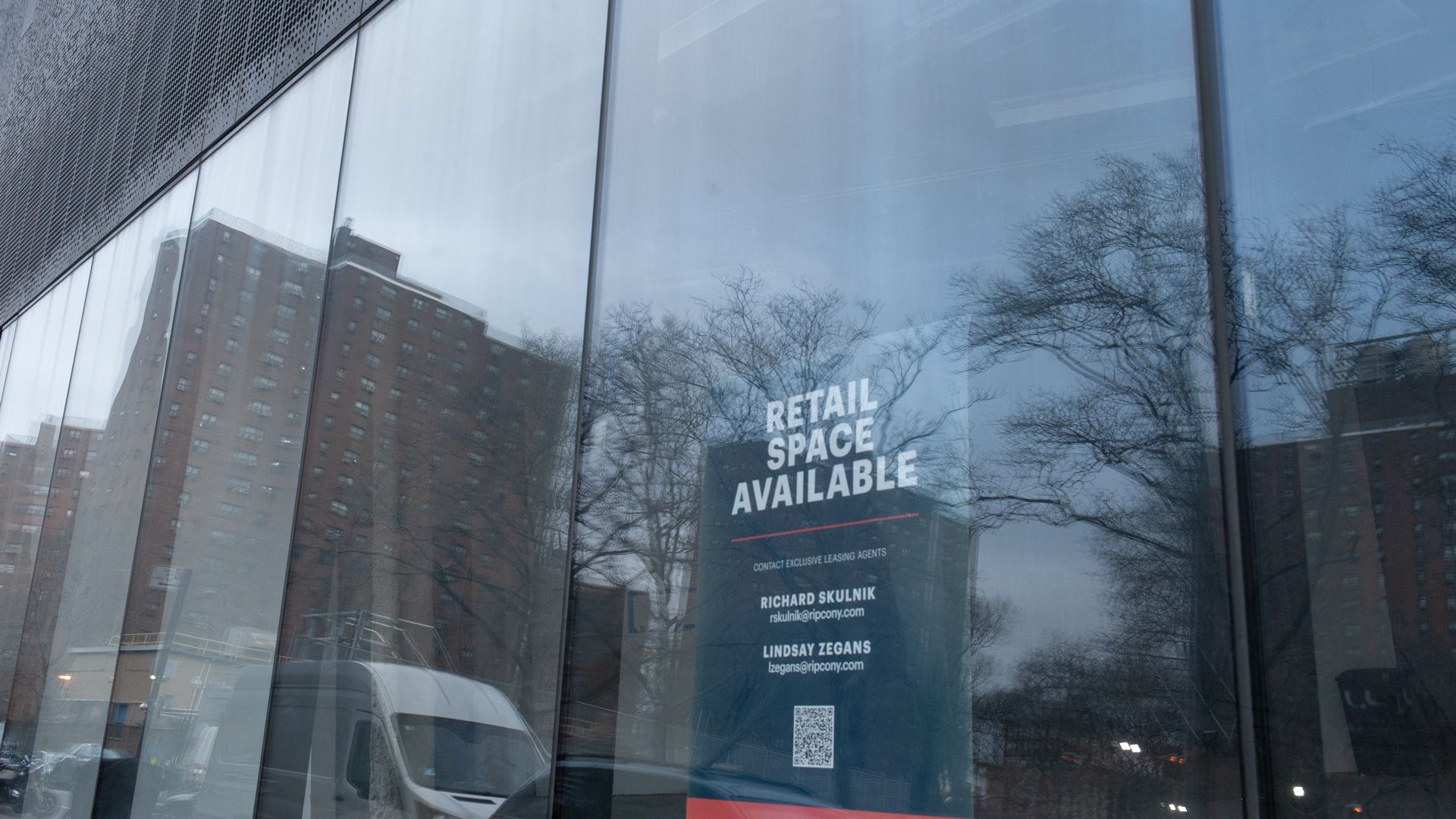 A photograph of a "retail space available" sign in the window of a new development building. Older buildings surrounding the property are reflected in the same window.
