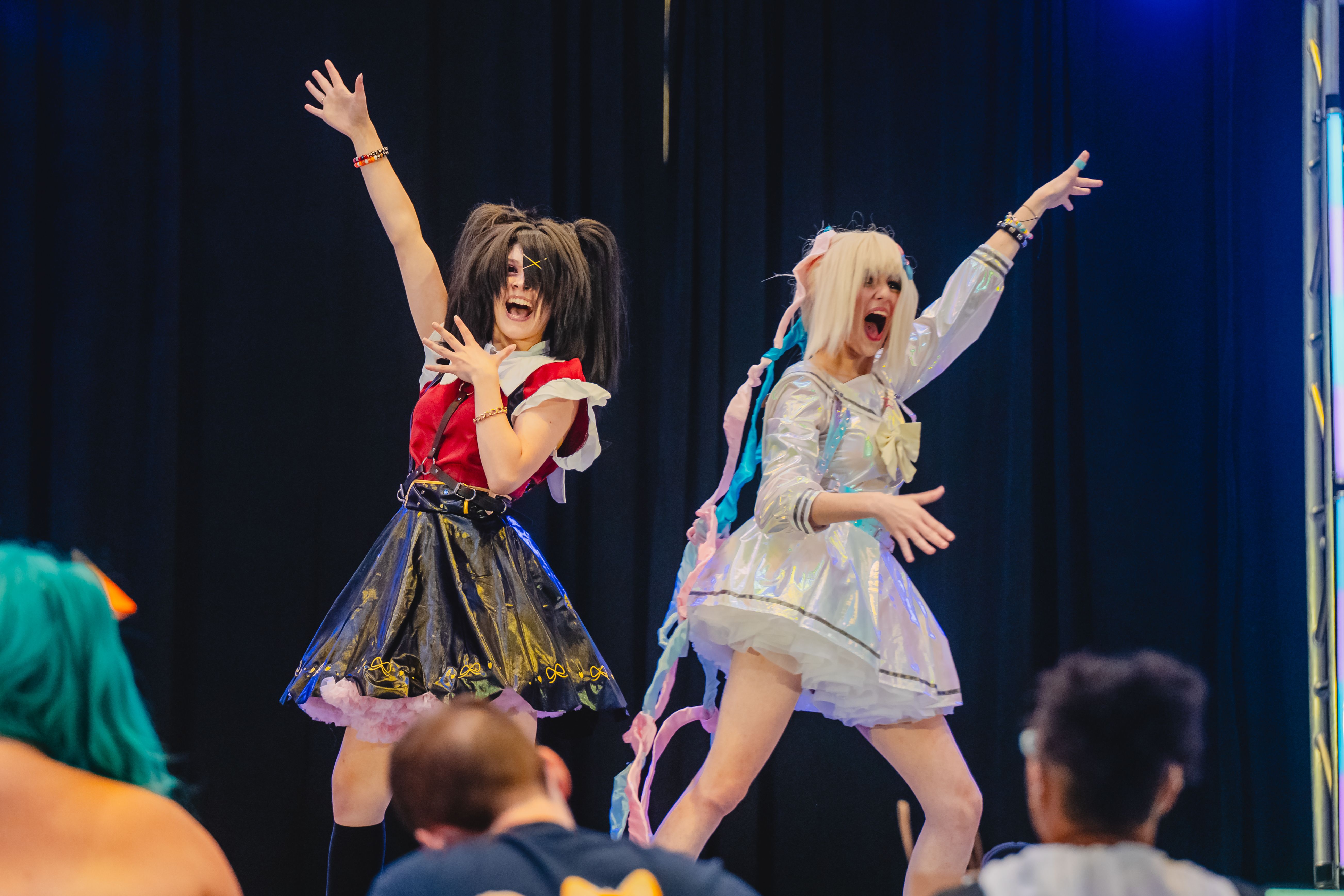 Two energetic cosplayers on stage, one in a red top with black skirt and dark pigtails, the other in a shiny white dress with pastel ribbons in blonde hair, performing dramatically before an audience.