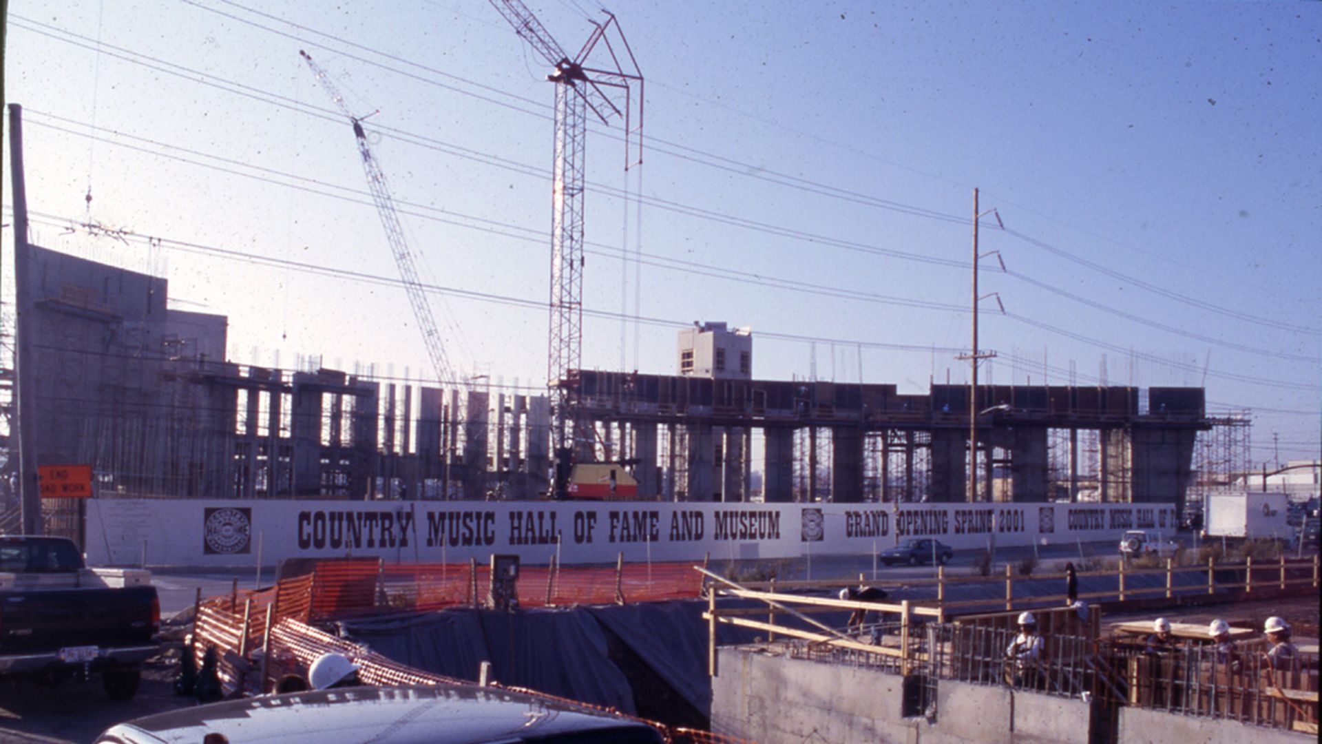 Construction site with cranes and scaffolding for the Country Music Hall of Fame and Museum; banner reads Grand Opening Spring 2001; blue sky, power lines, orange fencing, and parked cars in the foreground.