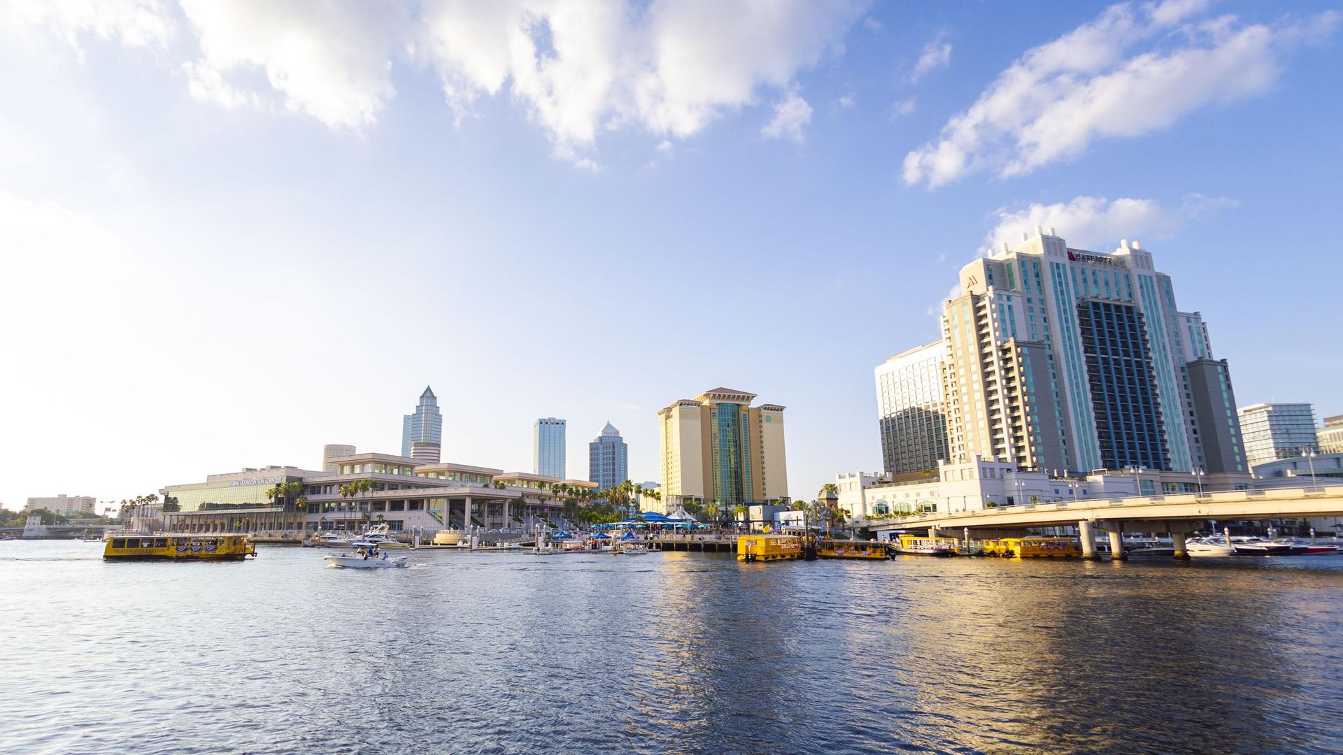 Waterfront cityscape with modern buildings, boats, and yellow water taxis on a sunny day under a blue sky with scattered white clouds.