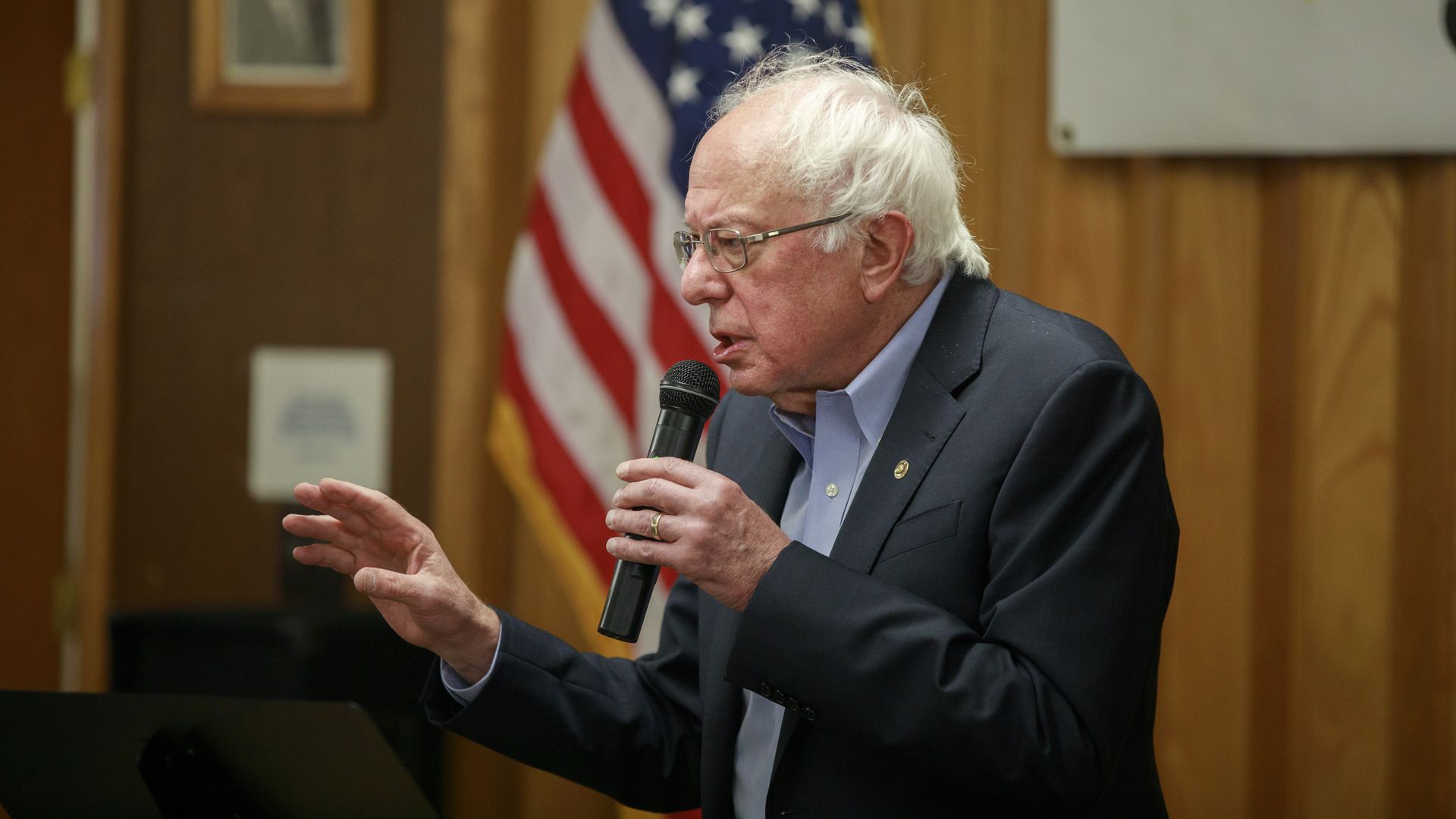 In this image, Bernie Sanders speaks into a microphone with an American flag behind him.