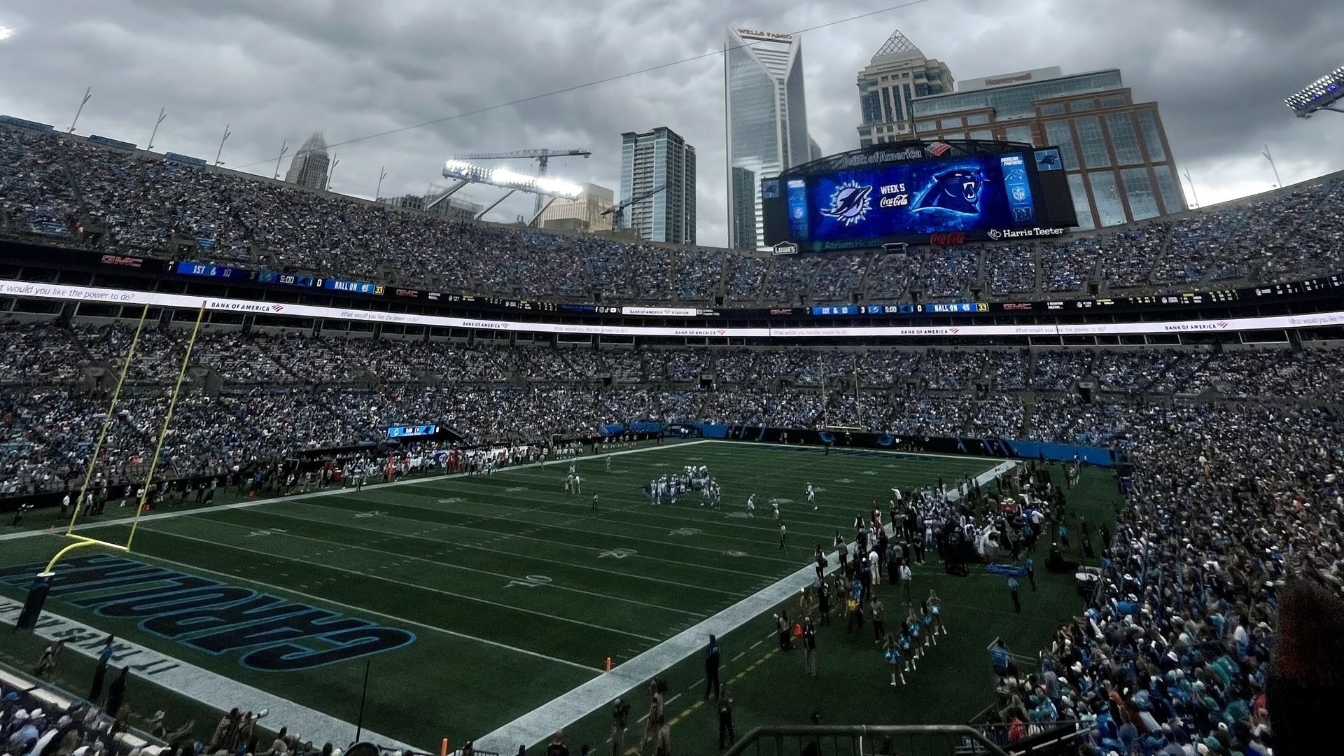 Overcast day at a packed football stadium; a large video board displays Panthers graphics while a city skyline looms behind the stands and players line the green field.