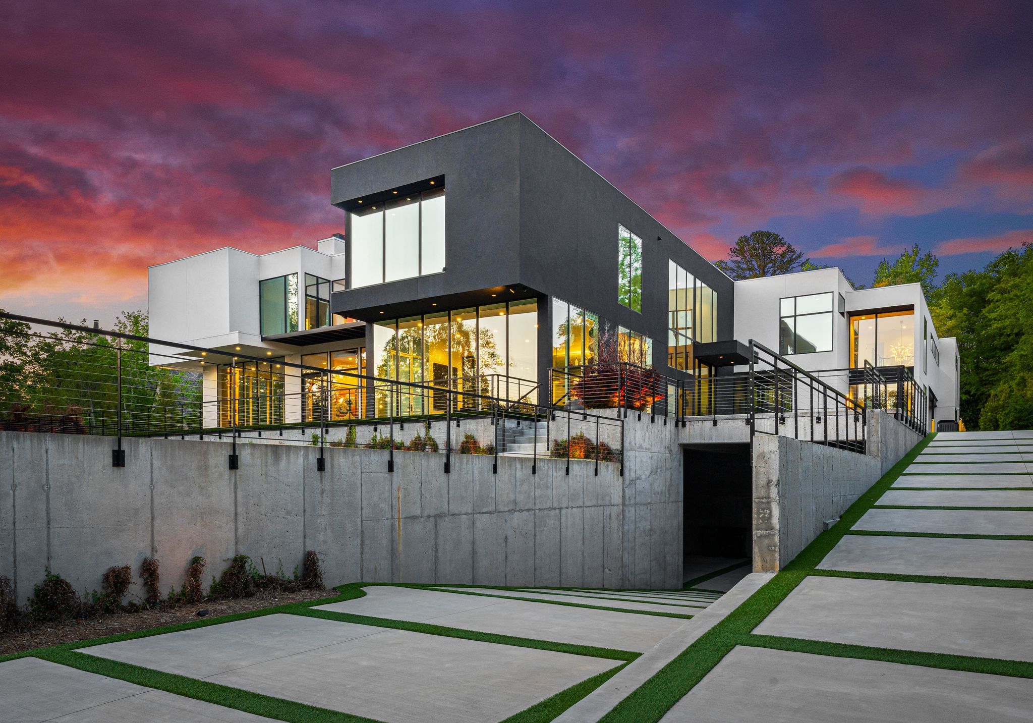 Modern multi-level house with large floor-to-ceiling windows, black and white exterior, concrete driveway with green stripes, surrounded by trees, under a colorful sunset sky.