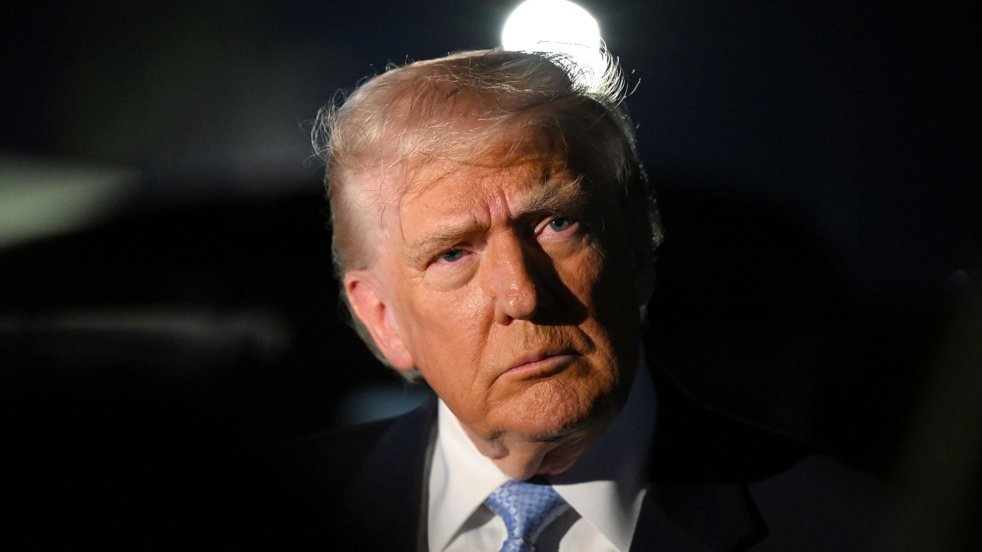President Trump, wearing a navy jacket with a US flag pin at the top of his left lapel, white shirt and blue tie, stares to his left.