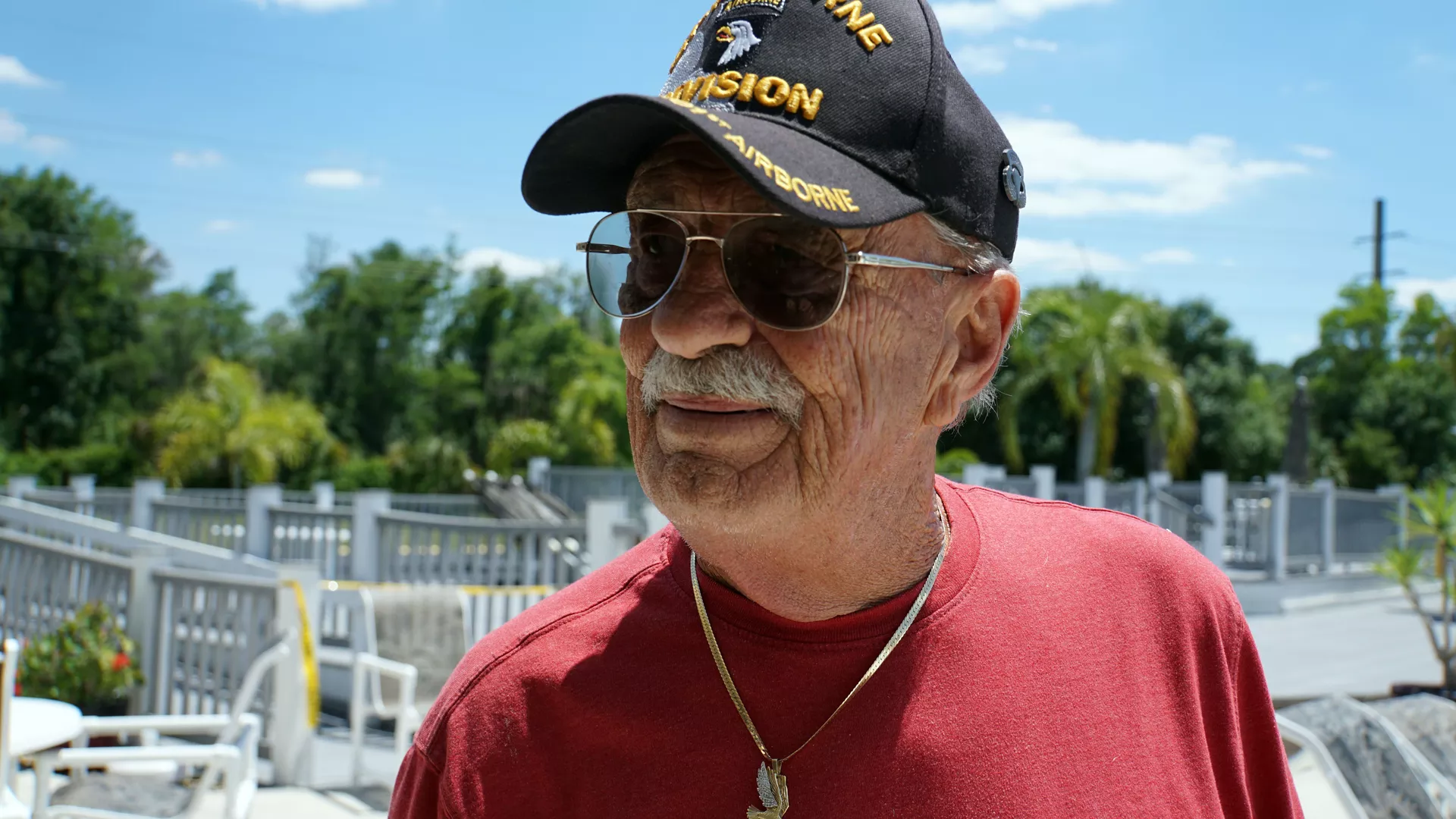 An older man with a mustache and a red t-shirt wearing a hat and glasses