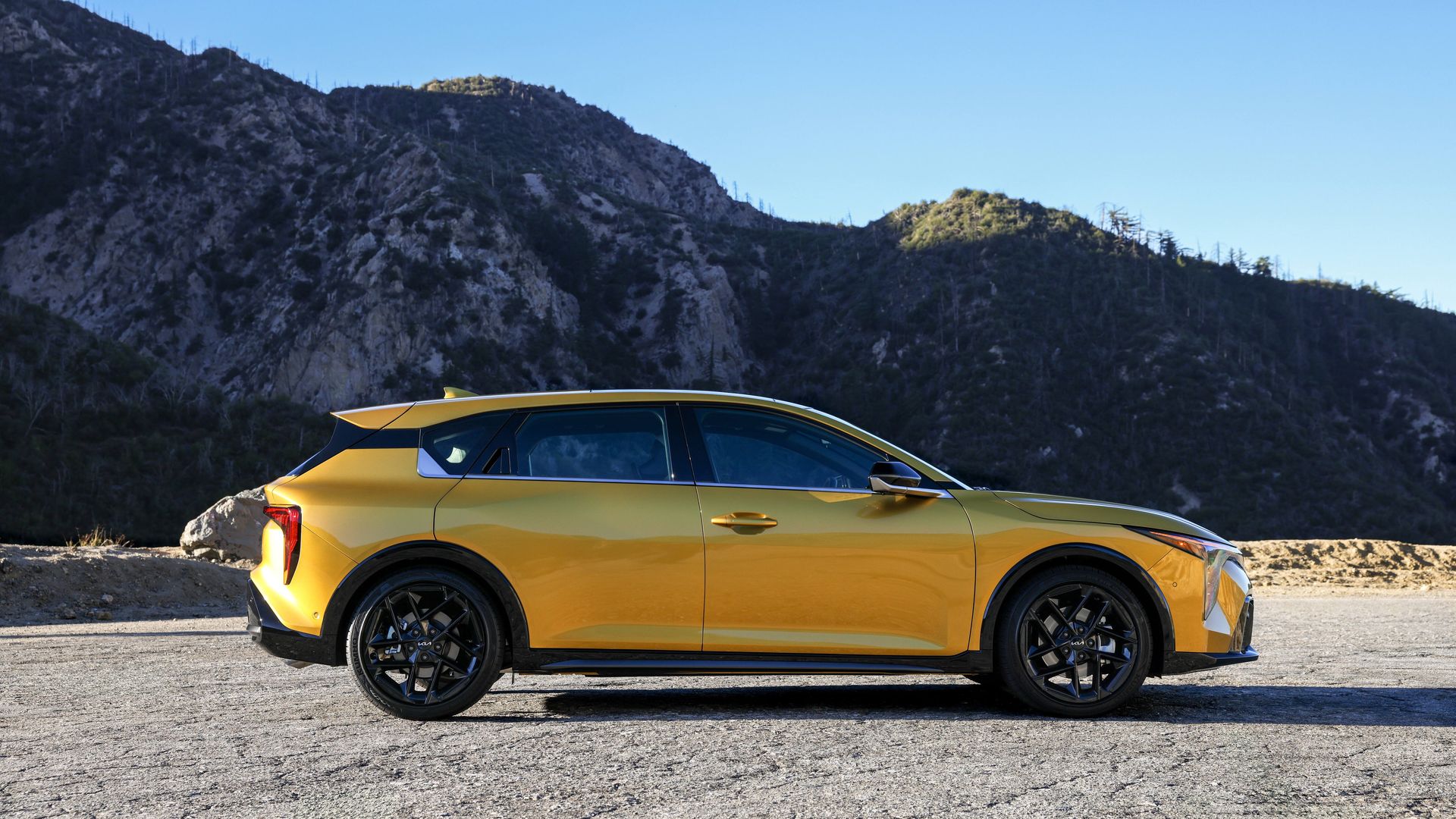 Bright yellow Kia 4 hatchback with black wheels sits on a gravel road, with rugged, tree-sparse mountains and a clear blue sky behind.