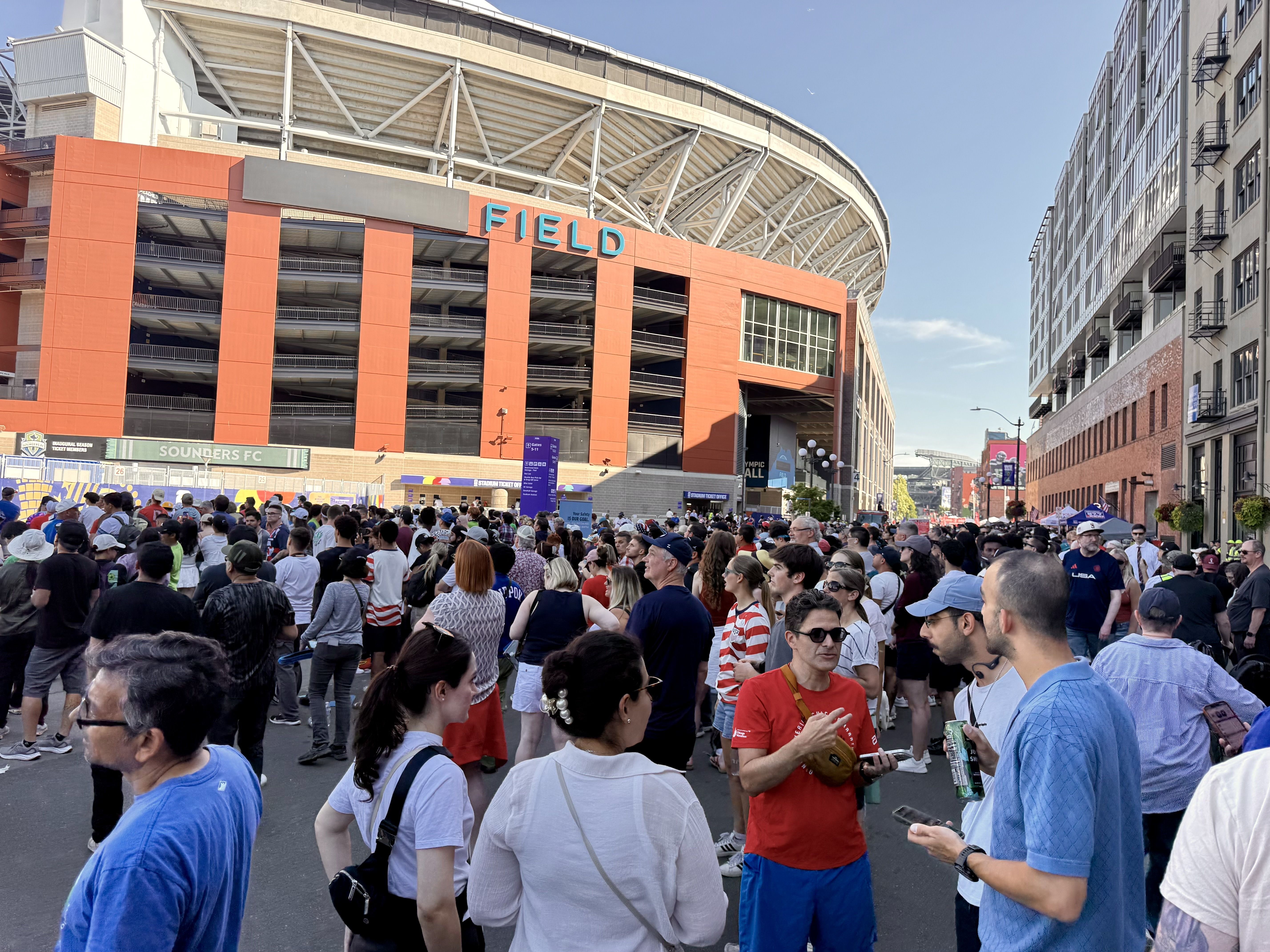 Large stadium with an orange exterior and turquoise "FIELD" sign looms over a crowded street; white steel roof trusses stretch above. Modern apartment buildings line the right, under a clear blue sky.