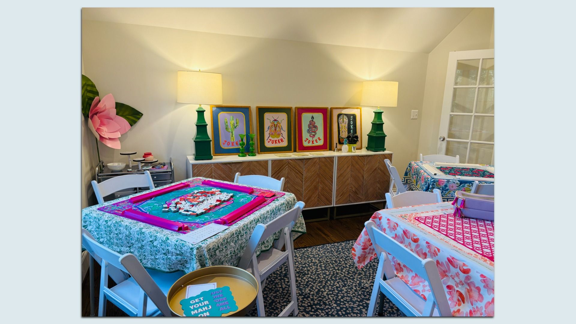 Game room with white folding chairs around three floral tables; mahjong-themed framed art on a sideboard, two green lamps, a pink flower decoration and a glass door on the right.