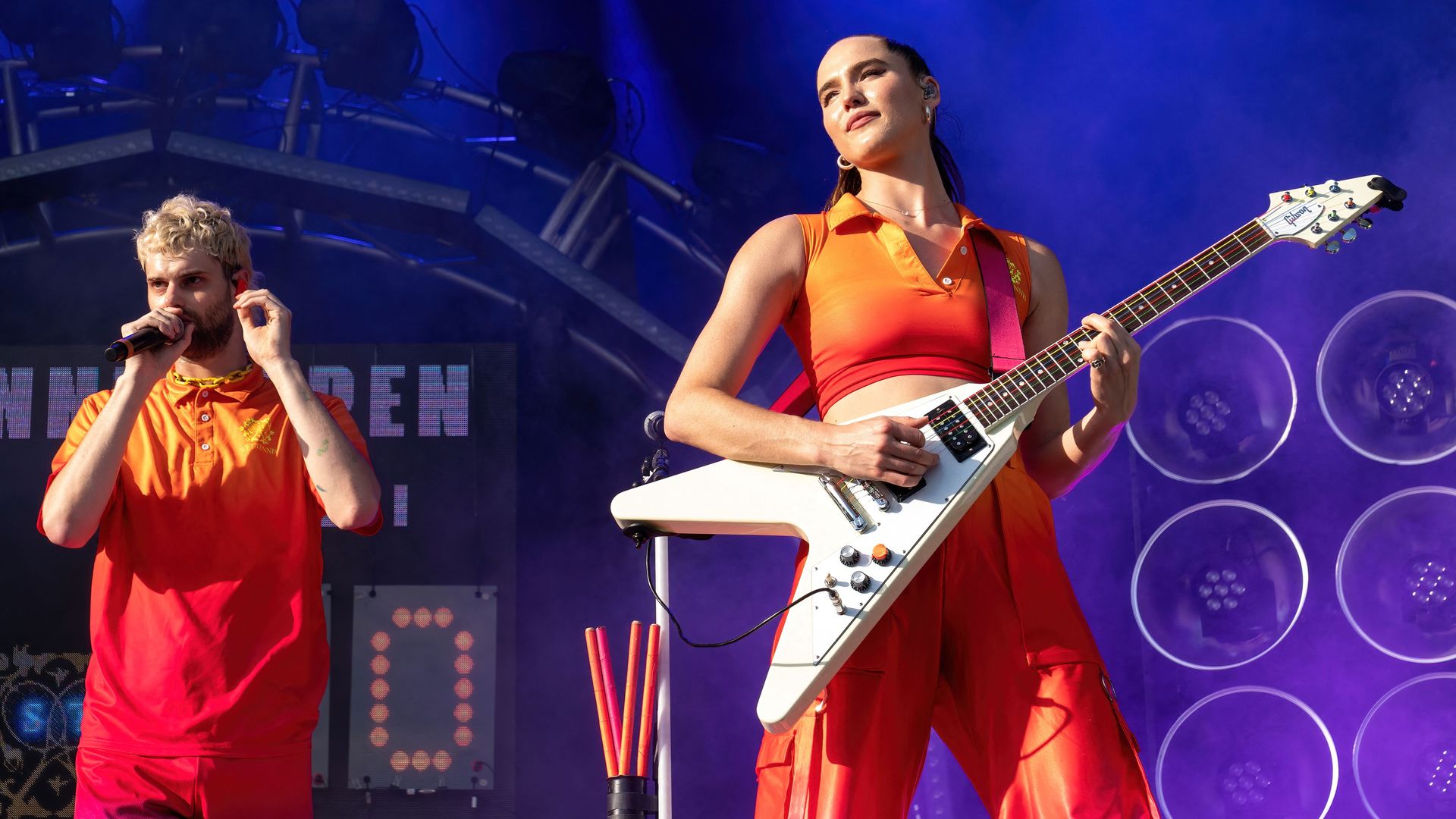 A guy with a microphone and woman with a v-shaped guitar wear bright orange outfits on stage 