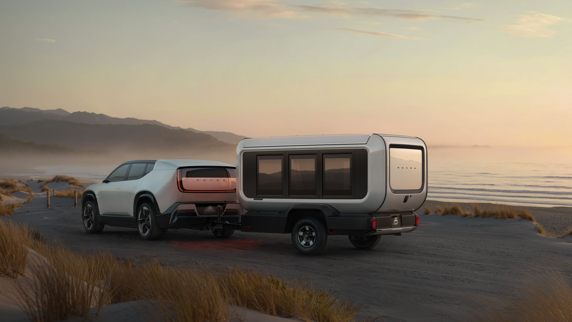 White Honda electric SUV towing a sleek, modern camper trailer on a sandy beach at sunset with mountains and ocean waves in the background.