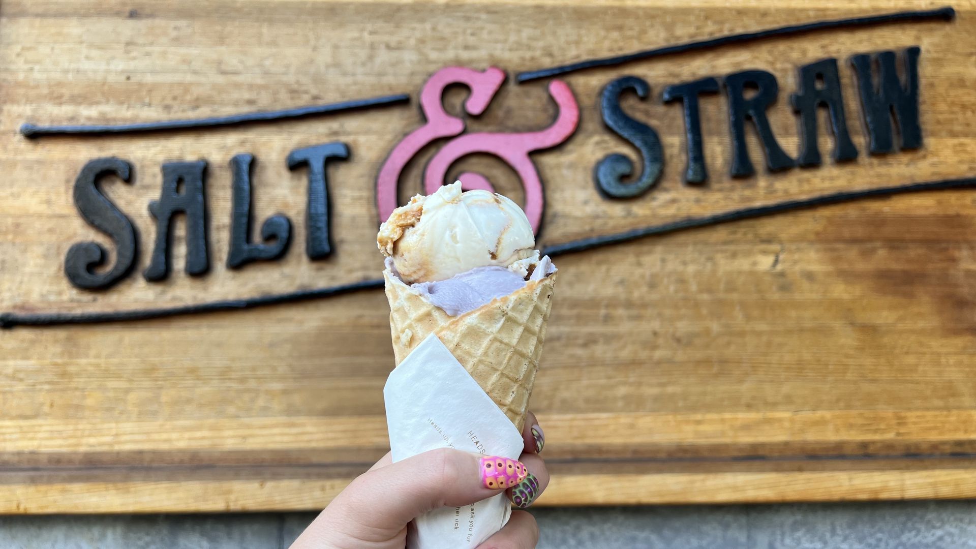 An image of a hand holding up an ice cream cone with the words Salt and Straw on a wooden board in the background.