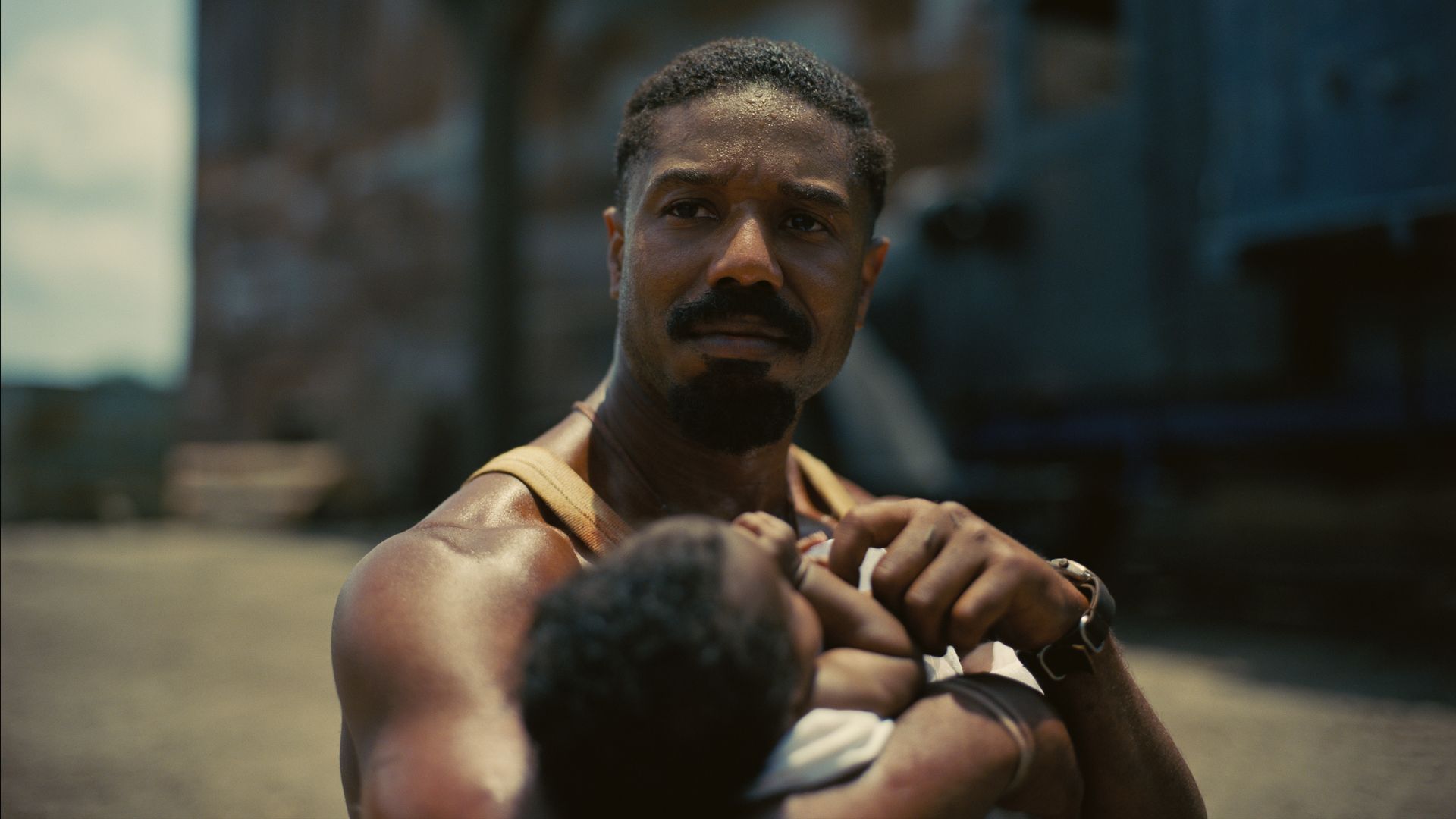 Man with a beard and mustache wearing a sleeveless tan shirt, holding a baby close, outdoors with blurred industrial background.