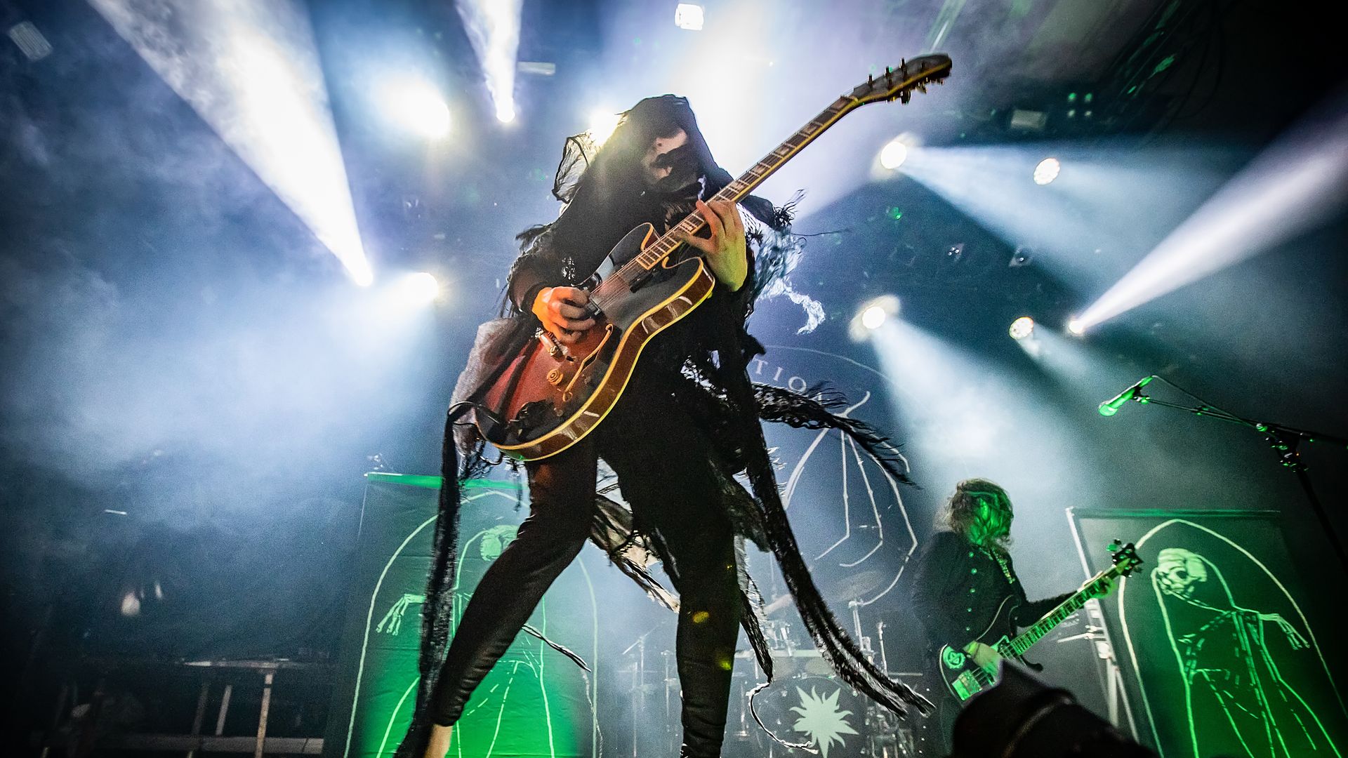 A heavy metal musician dressed in black performs on stage with a guitar