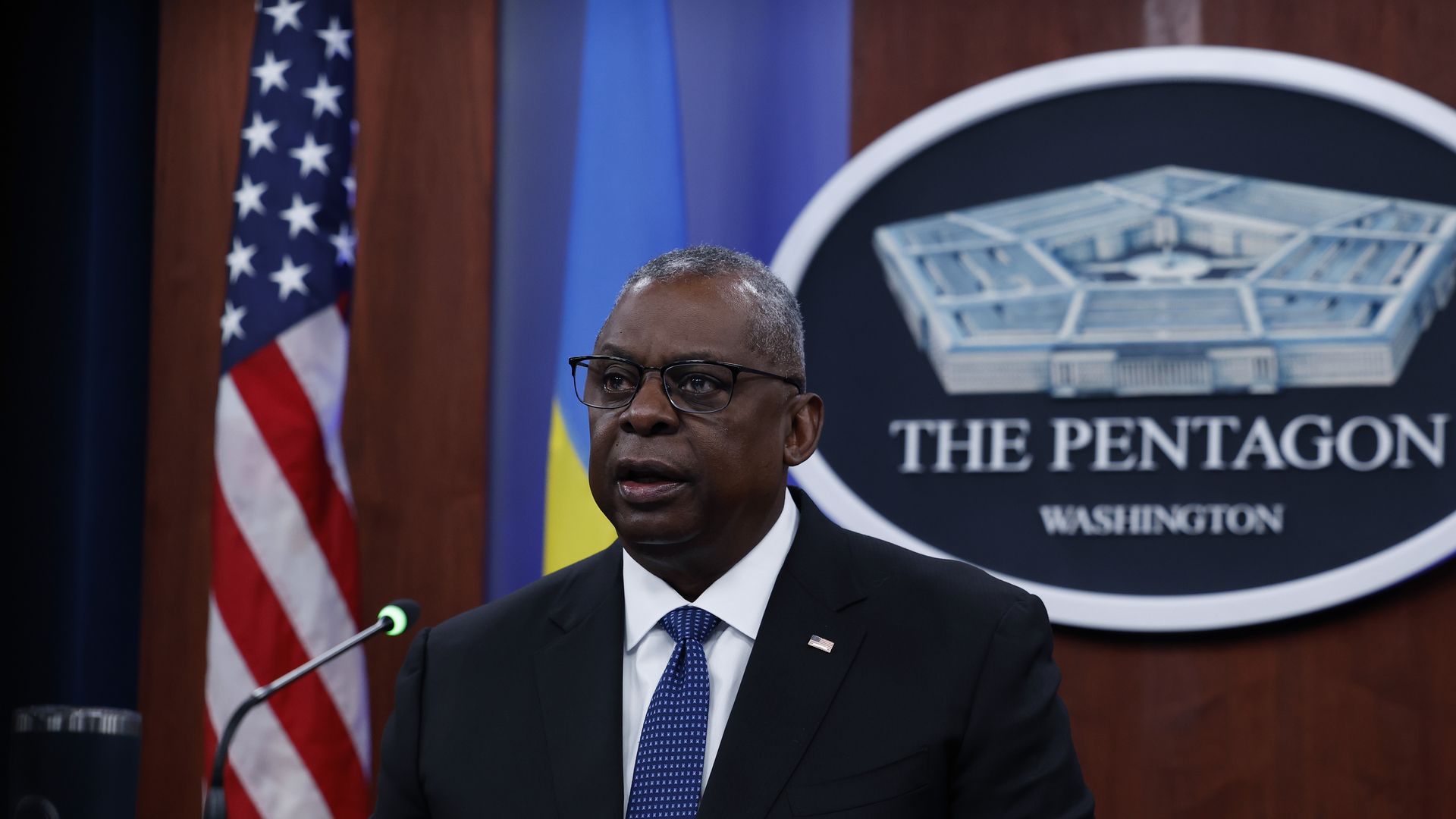 Lloyd Austin speaks into a microphone with an image of the Pentagon behind him and an American flag off to the side. 