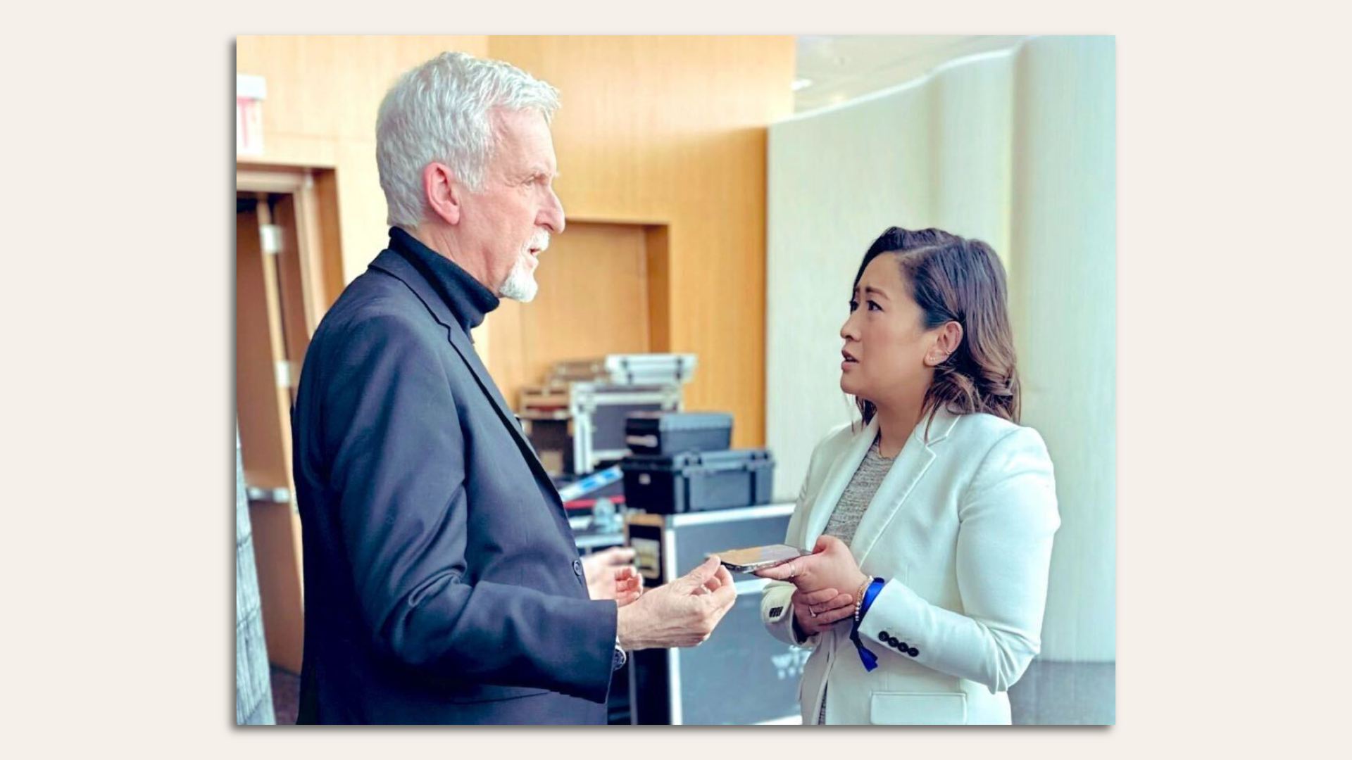 photo of a white haired man in a black turtleneck and blazer talking to a younger woman in a white blazer who is holding a phone